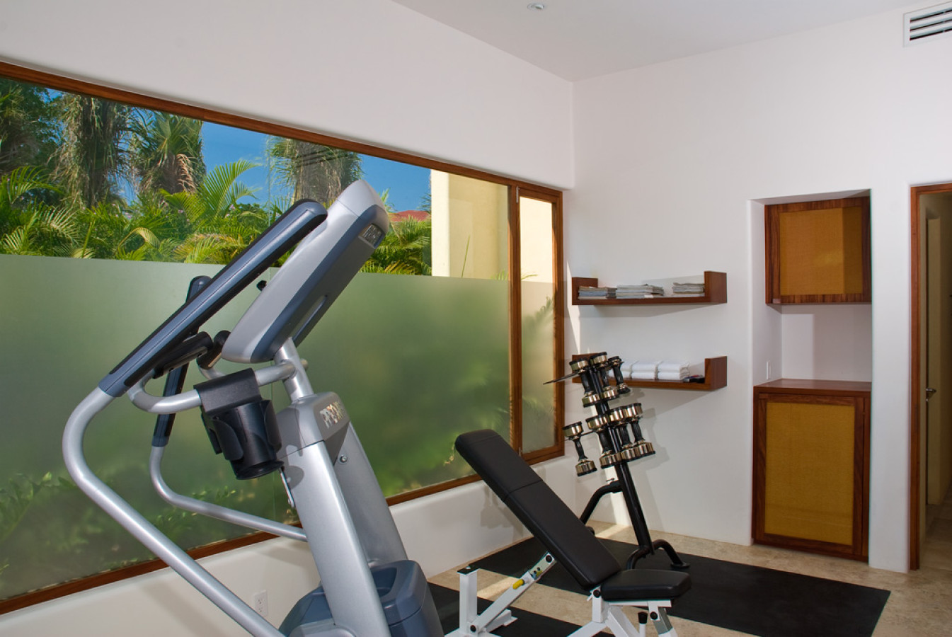 Fitness room with elliptical trainer, adjustable weight bench, free-weight rack, frosted glass garden window, walnut shelving with rolled towels, and dark wood doors, Estate Jaguar, Ranchos Estates, Punta Mita.
