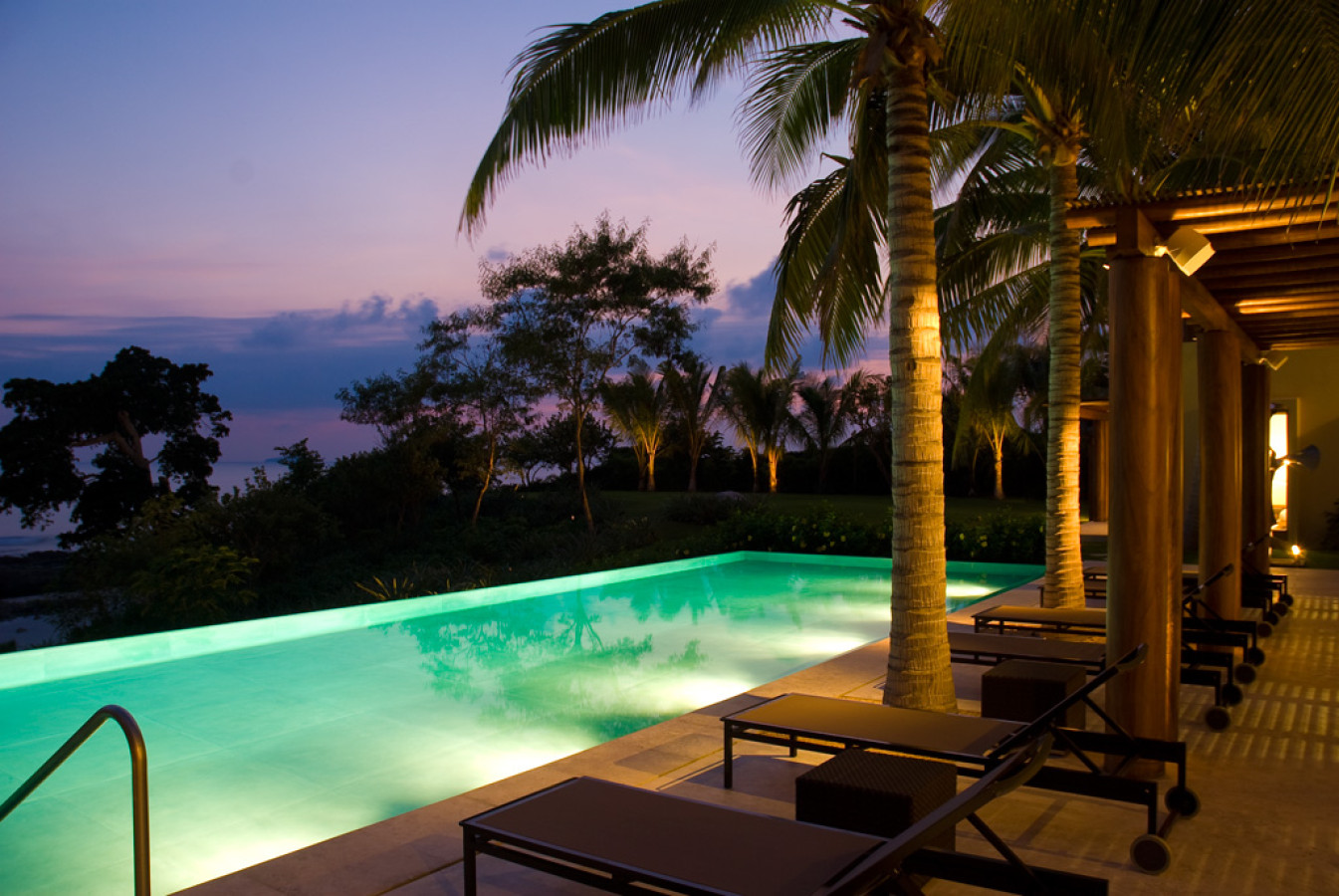 Infinity pool glowing teal under a pink dusk sky, lounge chairs beneath palm trees, flat-roof pavilion at right, Estate Jaguar, Ranchos Estates, Punta Mita.