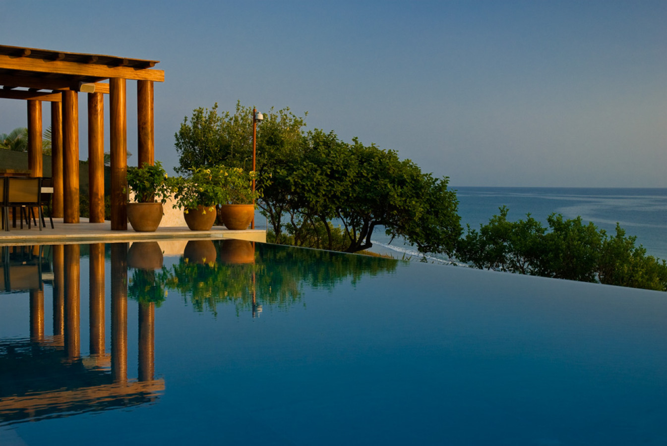 Still infinity pool at golden hour reflecting timber pergola columns and terracotta planters, Pacific horizon beyond, Estate Jaguar, Ranchos Estates, Punta Mita.