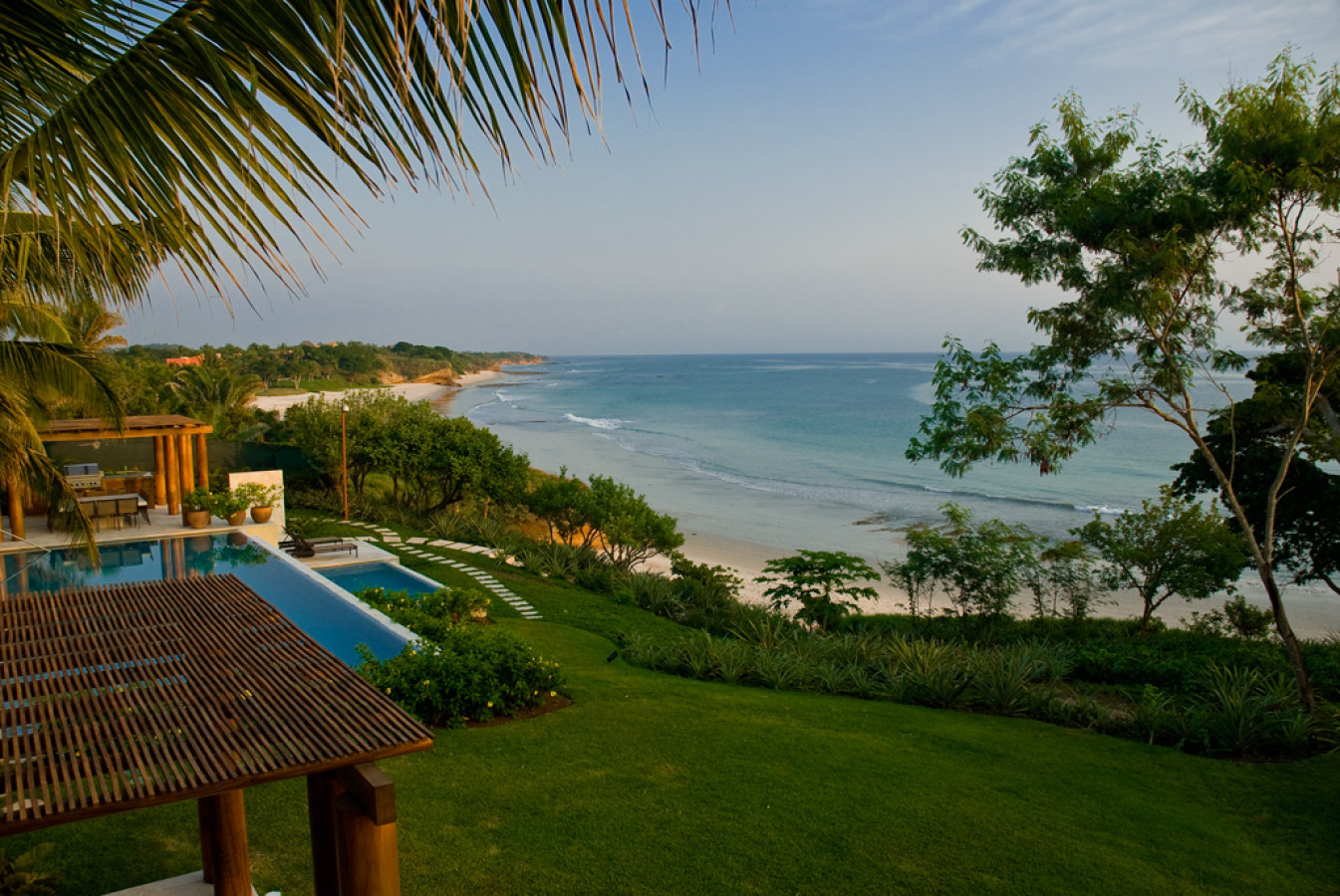 Terraced lawn and lap pool descending to a wide arc of white-sand beach, timber pergola in foreground, Estate Jaguar, Ranchos Estates, Punta Mita.