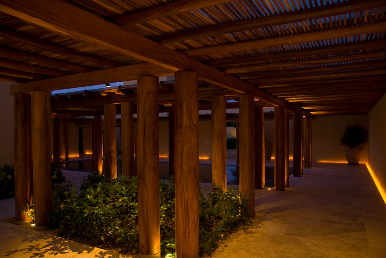Covered walkway at night with rows of heavy timber columns uplighted in amber, slatted wood ceiling, cream stucco walls, Estate Jaguar, Ranchos Estates, Punta Mita.