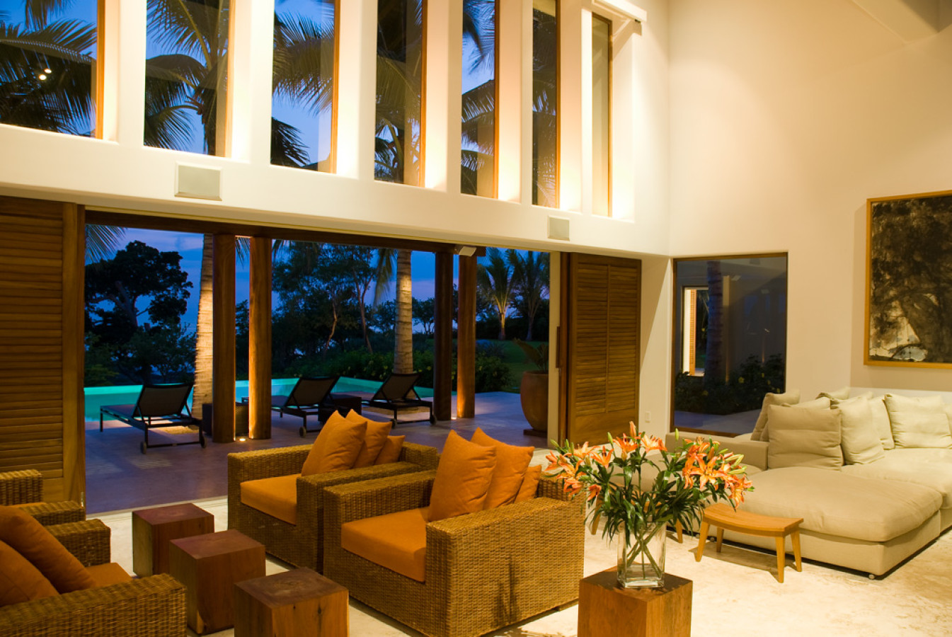 Living pavilion at dusk with double-height white walls, rattan seating with terracotta cushions, open louvered doors to the pool terrace and palm garden, Estate Jaguar, Ranchos Estates, Punta Mita.