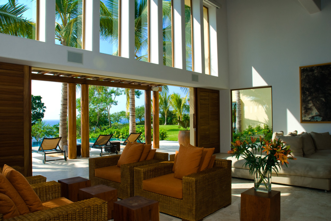 Sunlit living pavilion with rattan armchairs, terracotta cushions, open folding doors to the pool and ocean, cut lilies on a side table, Estate Jaguar, Ranchos Estates, Punta Mita.