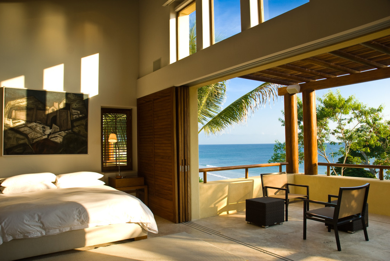 Primary suite with clerestory windows, dark-framed artwork, king bed, and folding doors open to a private terrace overlooking the Pacific, Estate Jaguar, Ranchos Estates, Punta Mita.
