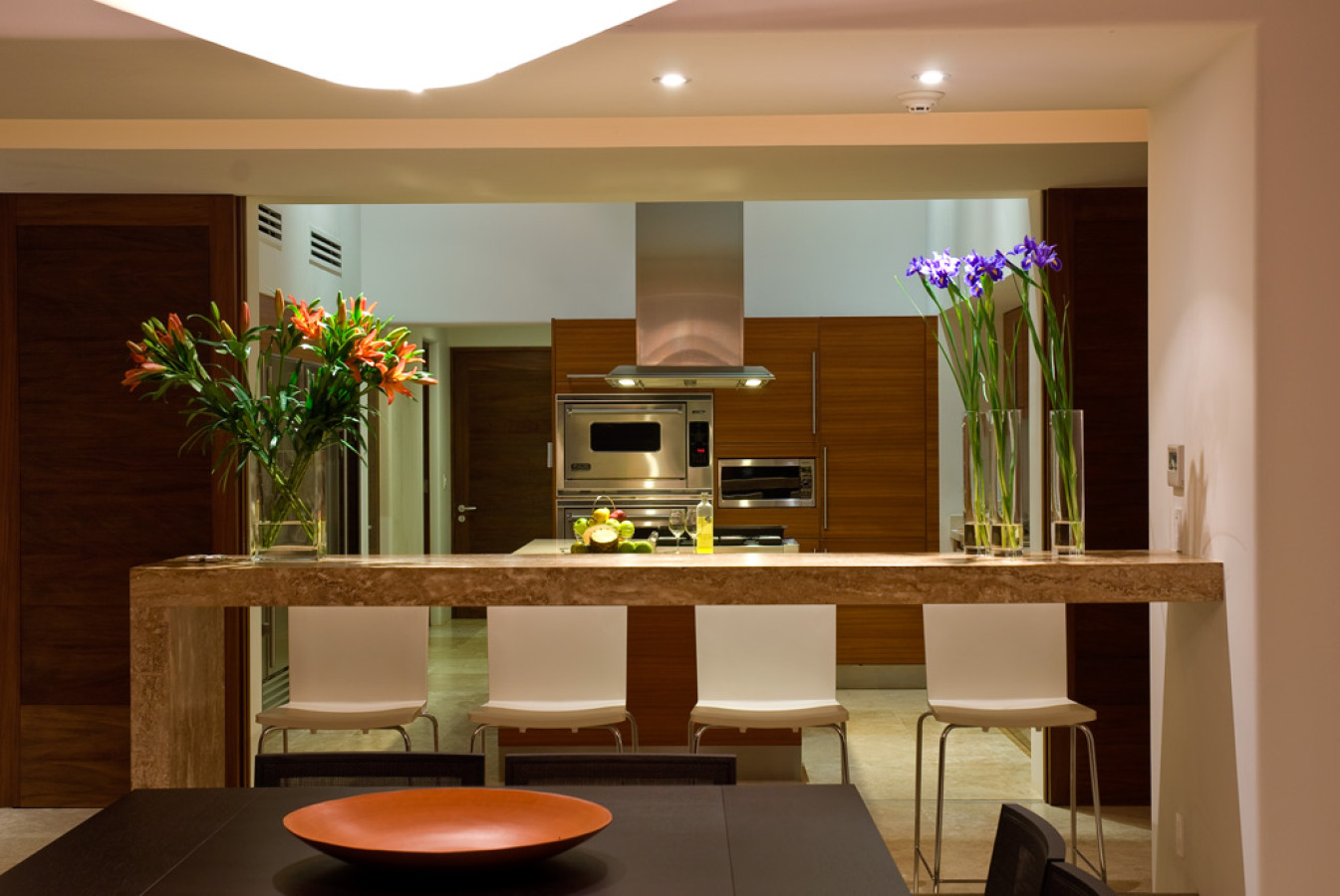 Kitchen pass-through bar with travertine counter, four white bar stools, stainless Viking range and hood, walnut cabinetry, cut lilies and irises, Estate Jaguar, Ranchos Estates, Punta Mita.