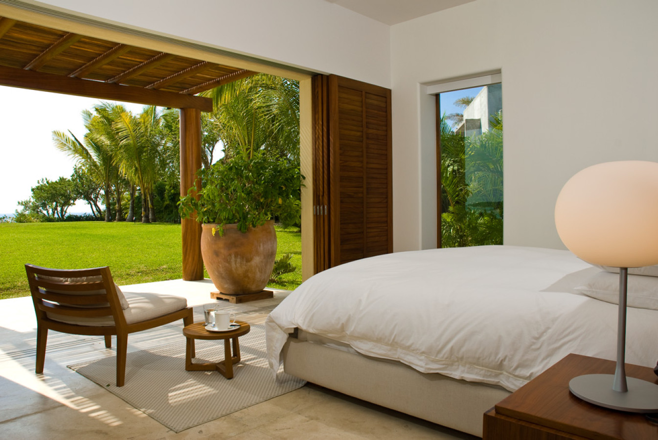 Guest suite with platform bed, globe bedside lamp, louvered shutters open to a shaded terrace with a large terracotta planter and manicured lawn, Estate Jaguar, Ranchos Estates, Punta Mita.