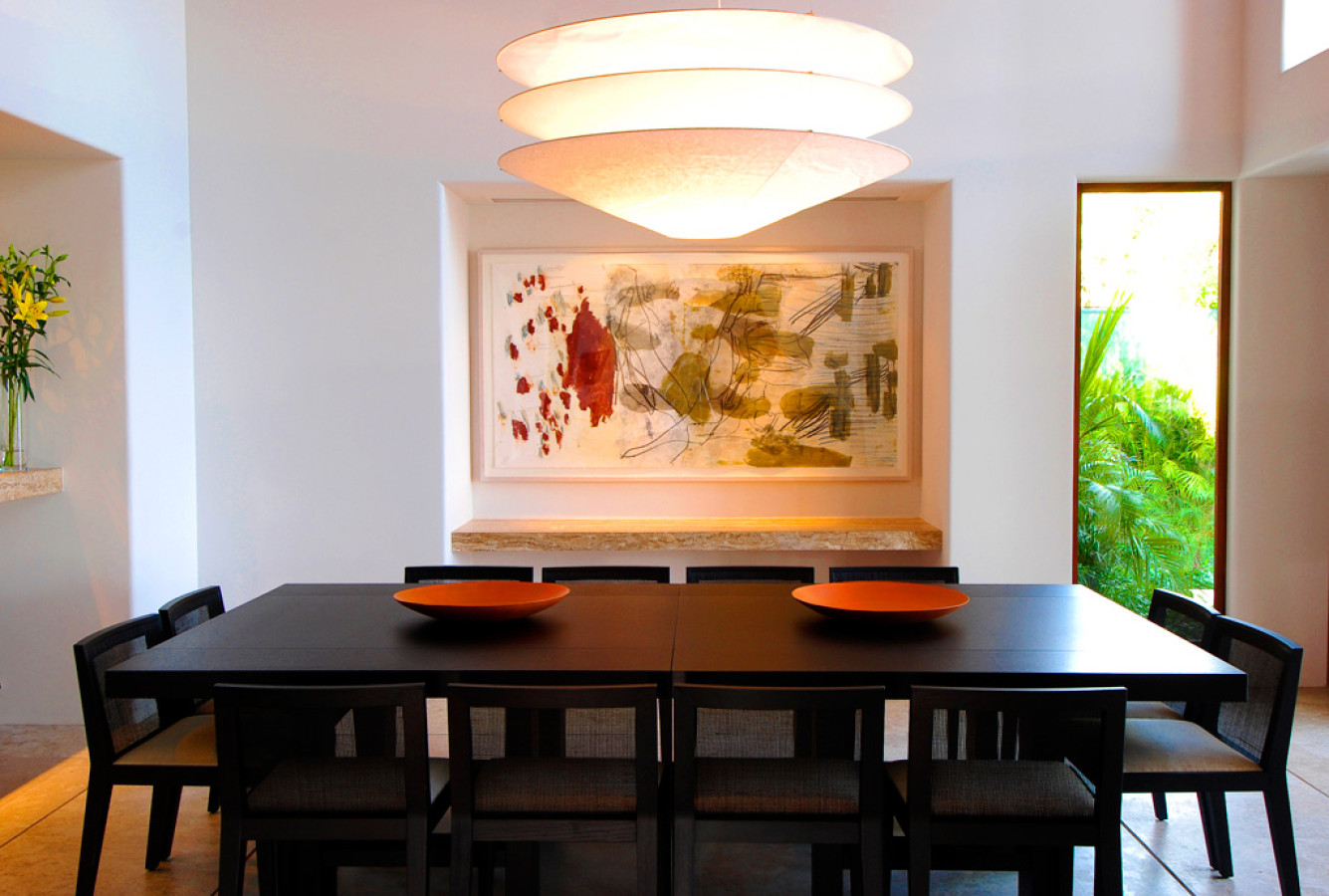 Formal dining room with a dark wood table for eight, stacked disc pendant, large abstract artwork on a white plaster wall, and a garden glimpse at right, Estate Jaguar, Ranchos Estates, Punta Mita.
