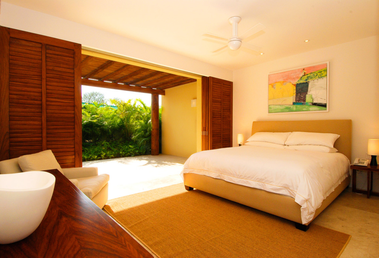 Guest suite with upholstered queen bed, sisal rug, ceiling fan, colorful framed painting, and louvered shutters open to a garden terrace with a freestanding soaker tub in foreground, Estate Jaguar, Ranchos Estates, Punta Mita.