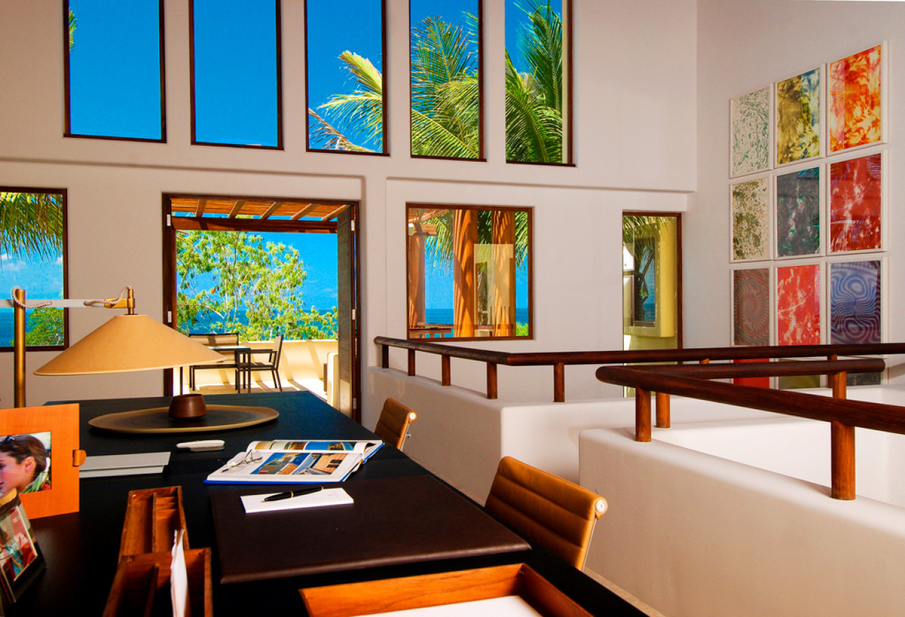 Upper-level study loft with desk, task lamp, open books, wood railing, tall clerestory windows framing palms and the Pacific, colourful grid artwork on white wall, Estate Jaguar, Ranchos Estates, Punta Mita.