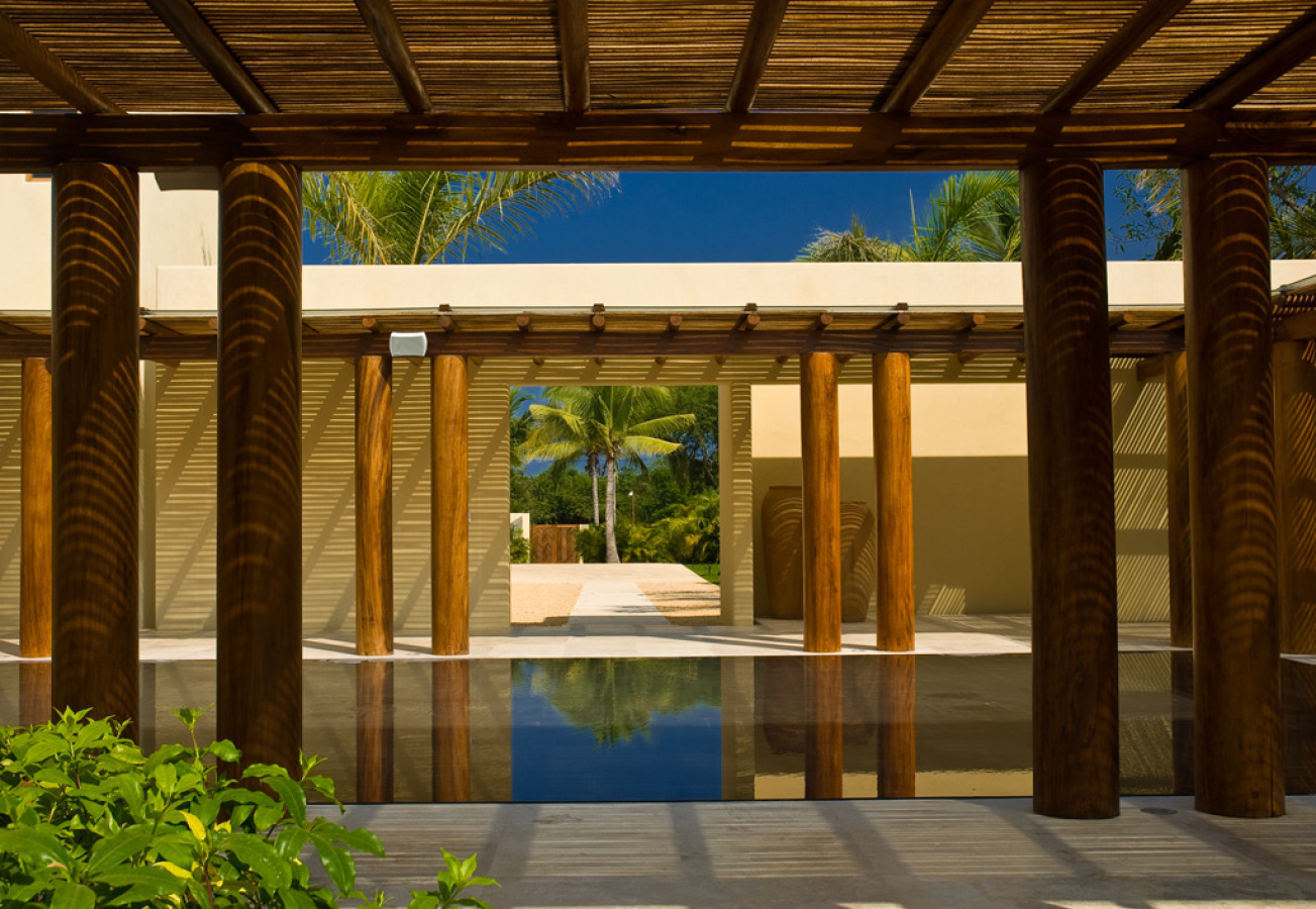 Arrival colonnade with heavy timber columns and slatted ceiling framing a long reflecting pool courtyard and a single palm beyond, Estate Jaguar, Ranchos Estates, Punta Mita.