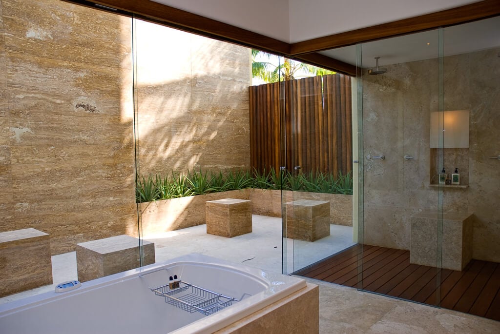 Primary ensuite with deep soaking tub, full-height travertine walls, glass rain shower on teak deck, and an open garden courtyard with bamboo screen beyond, Estate Jaguar, Ranchos Estates, Punta Mita.