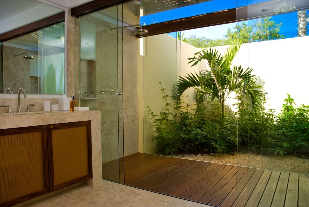 Ensuite bath with glass rain shower, teak deck floor, open-sky courtyard planted with a small palm and tropical shrubs, travertine vanity with wood-framed cabinets at left, Estate Jaguar, Ranchos Estates, Punta Mita.