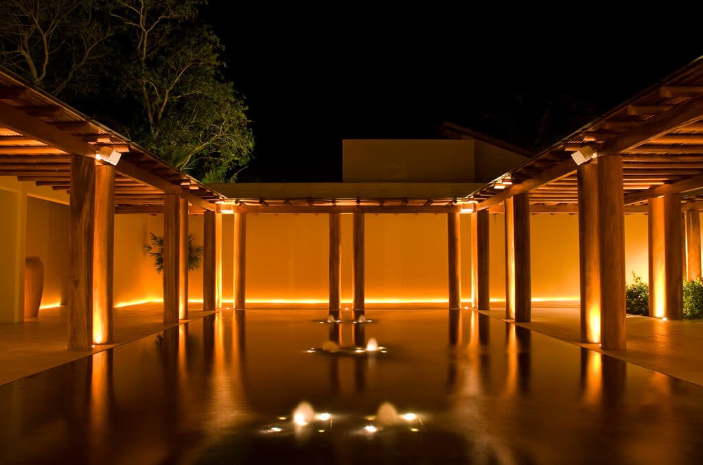 Entry courtyard reflecting pool at night with symmetrical timber colonnades, amber uplighting on cream stucco walls, and a small central fountain, Estate Jaguar, Ranchos Estates, Punta Mita.