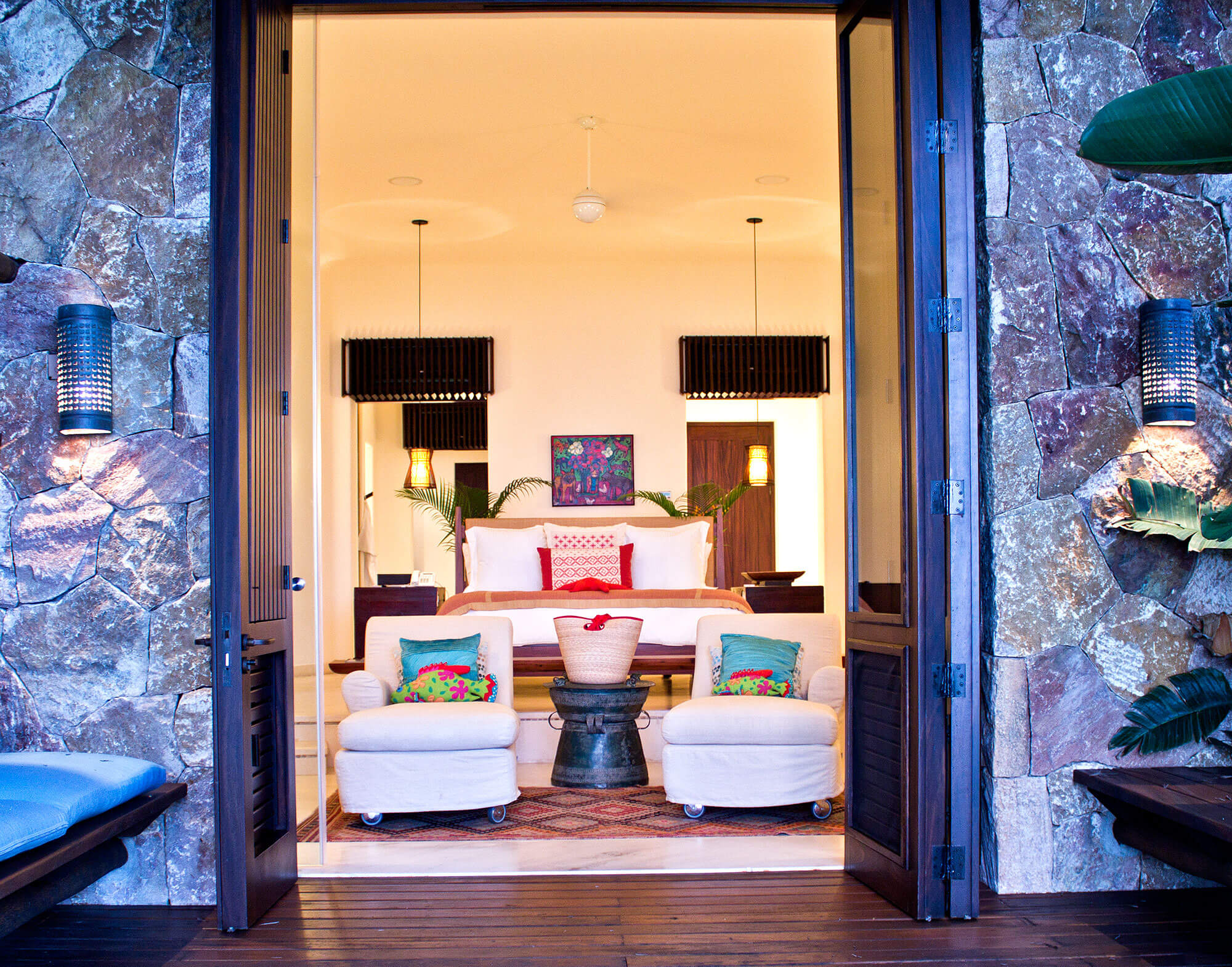 View through open glass doors into a suite interior — white lounge chairs with colorful pillows, king bed, pendant lights, and rough volcanic stone wall surrounding the entry terrace, Estate Venado, Ranchos Estates, Punta Mita.