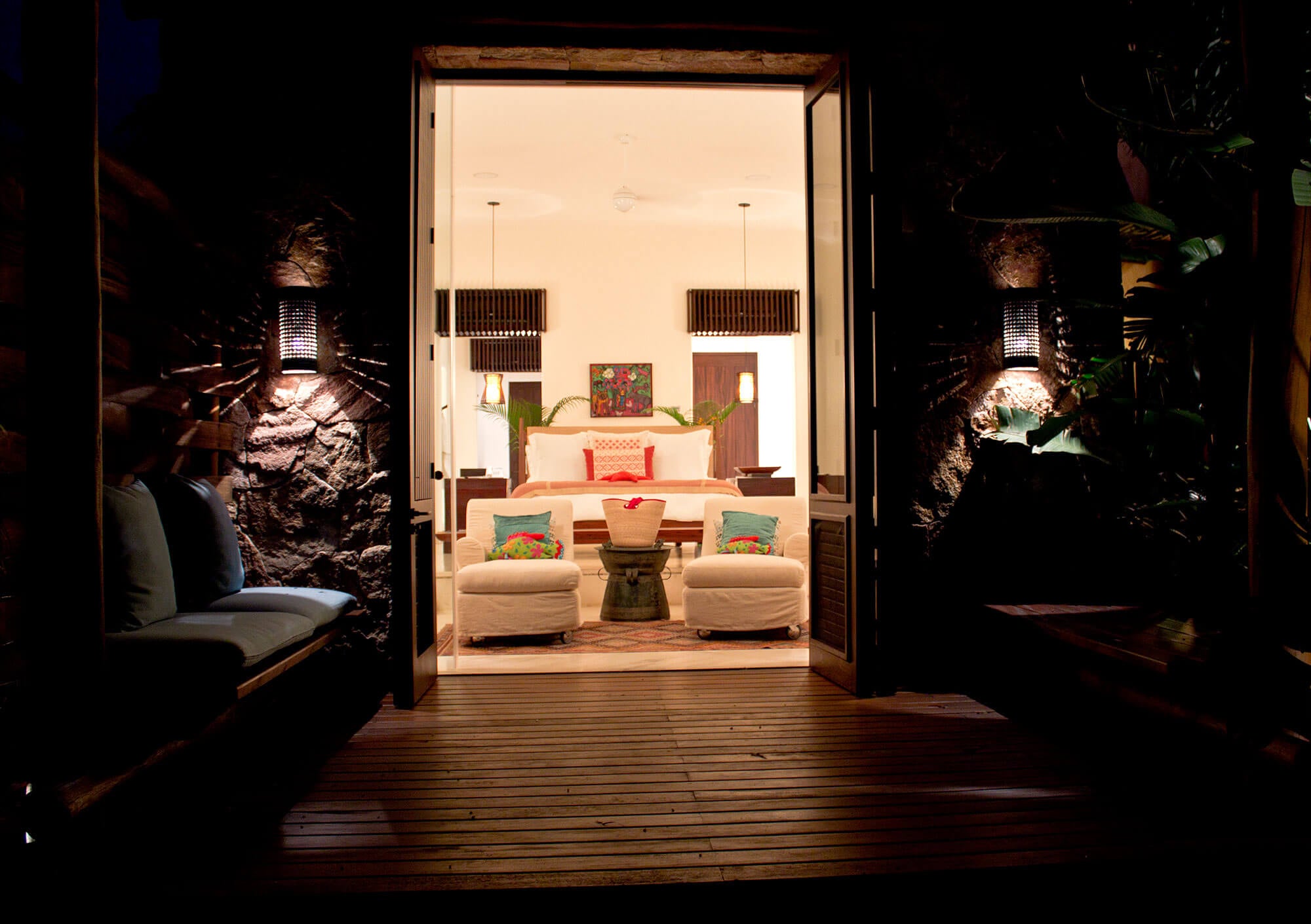 Night view from a teak deck through open suite doors — private hanging daybed at left, volcanic stone walls, lit bedroom interior with colorful accent pillows and king bed beyond, Estate Venado, Ranchos Estates, Punta Mita.