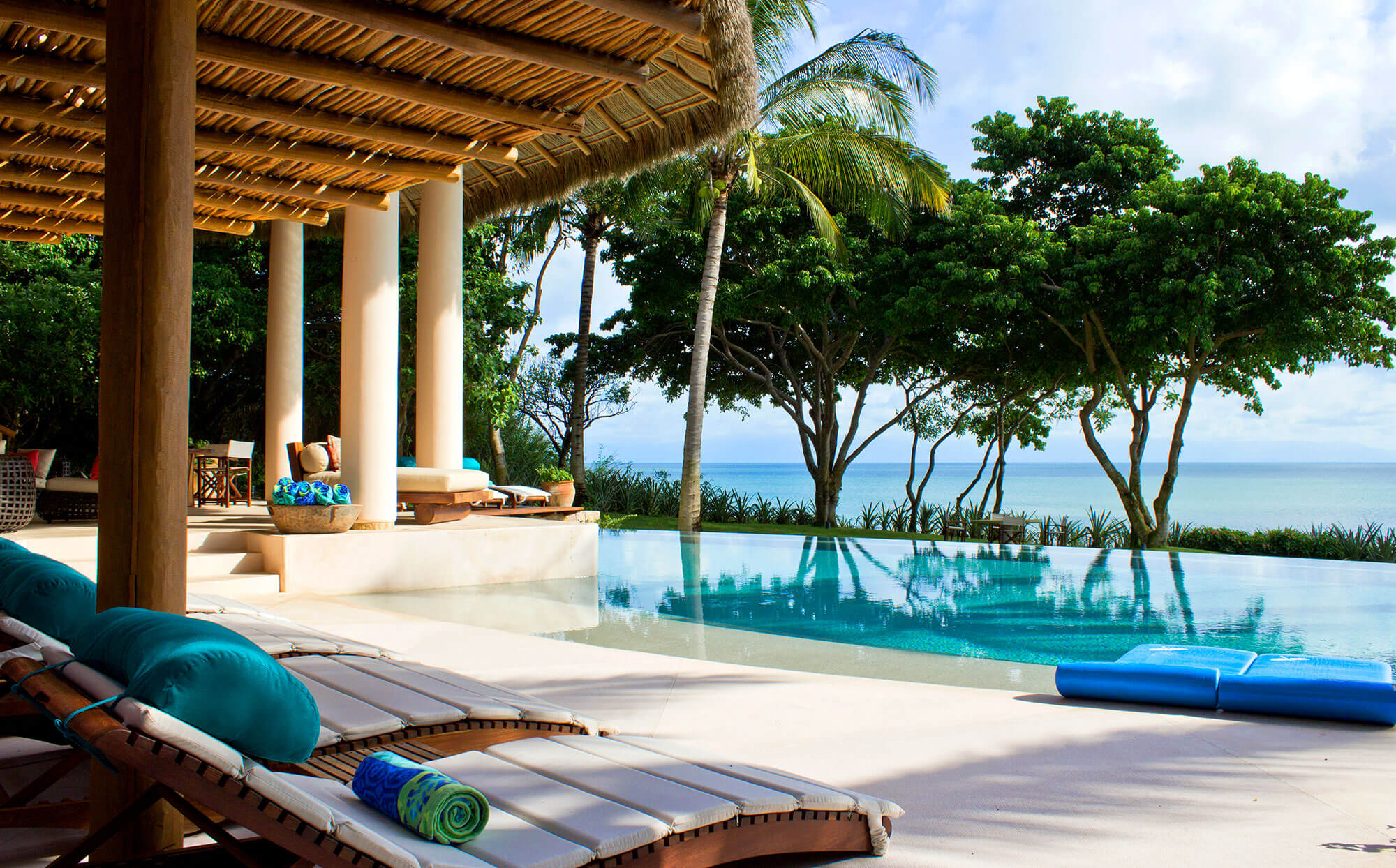 Infinity pool deck with teak sun loungers in the foreground, palapa-roofed colonnade at left, tall palms and tropical trees, and a broad Pacific ocean view along the pool edge, Estate Venado, Ranchos Estates, Punta Mita.