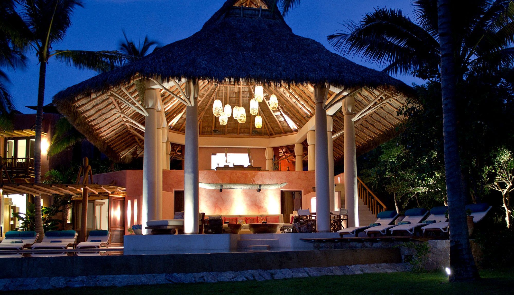 Night exterior of the estate's circular palapa-roofed living pavilion — woven pendant lanterns glowing inside the thatched canopy, round stucco columns, pool loungers and tropical garden flanking the open structure, Estate Venado, Ranchos Estates, Punta Mita.