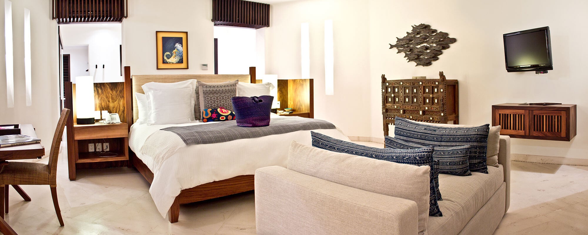 Primary suite interior with a king bed on warm wood headboard with integrated bedside lighting, cream sofa at the foot, carved wooden screen, school-of-fish metal wall sculpture, wall-mounted TV, and white marble floors, Estate Venado, Ranchos Estates, Punta Mita.