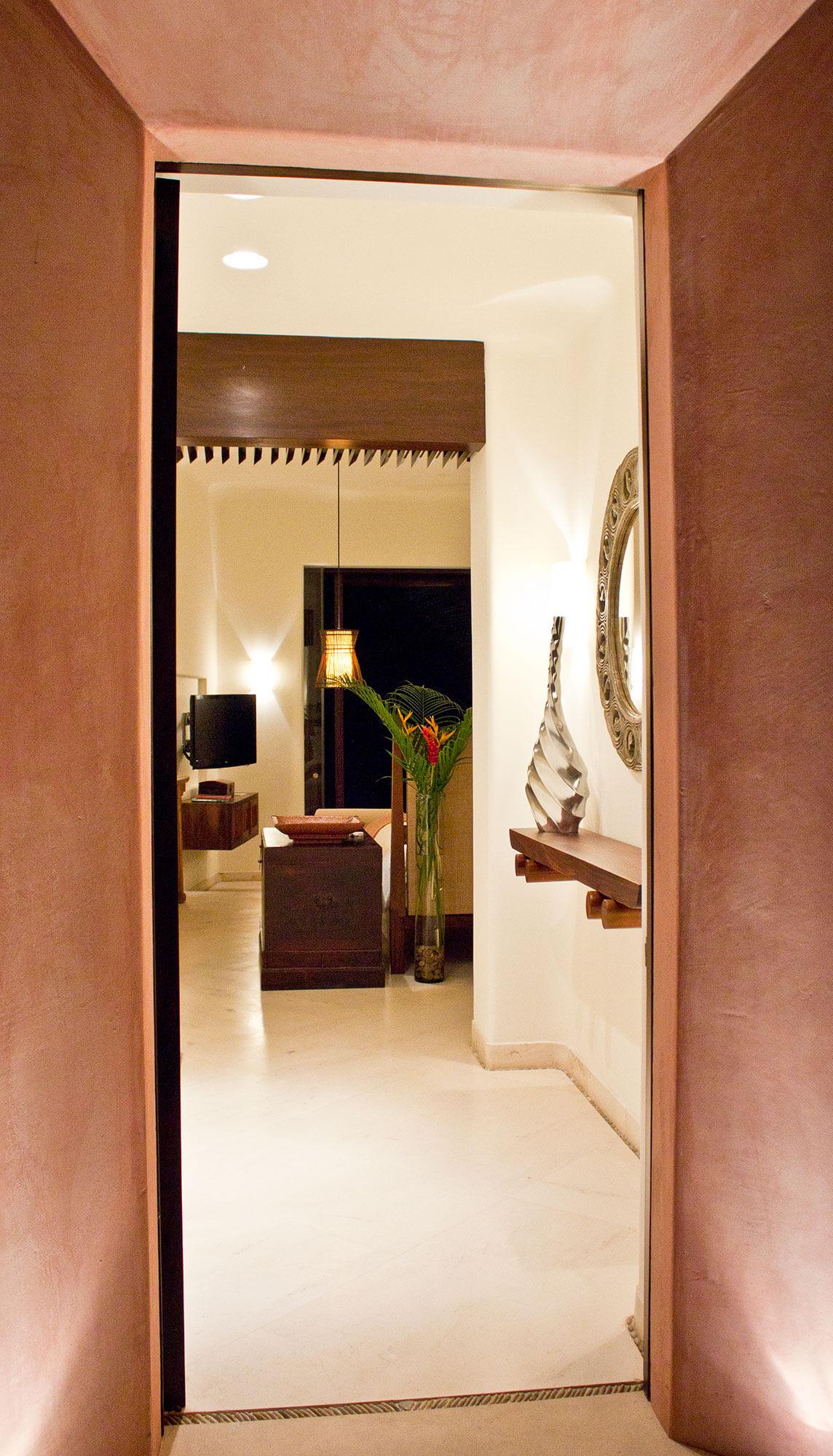 Narrow terracotta-plastered entry corridor with a glass door opening into a lit suite — ornate round mirror on a wood wall shelf, sculptural vase, tropical flower arrangement in a vase, and a pendant lantern beyond, Estate Venado, Ranchos Estates, Punta Mita.