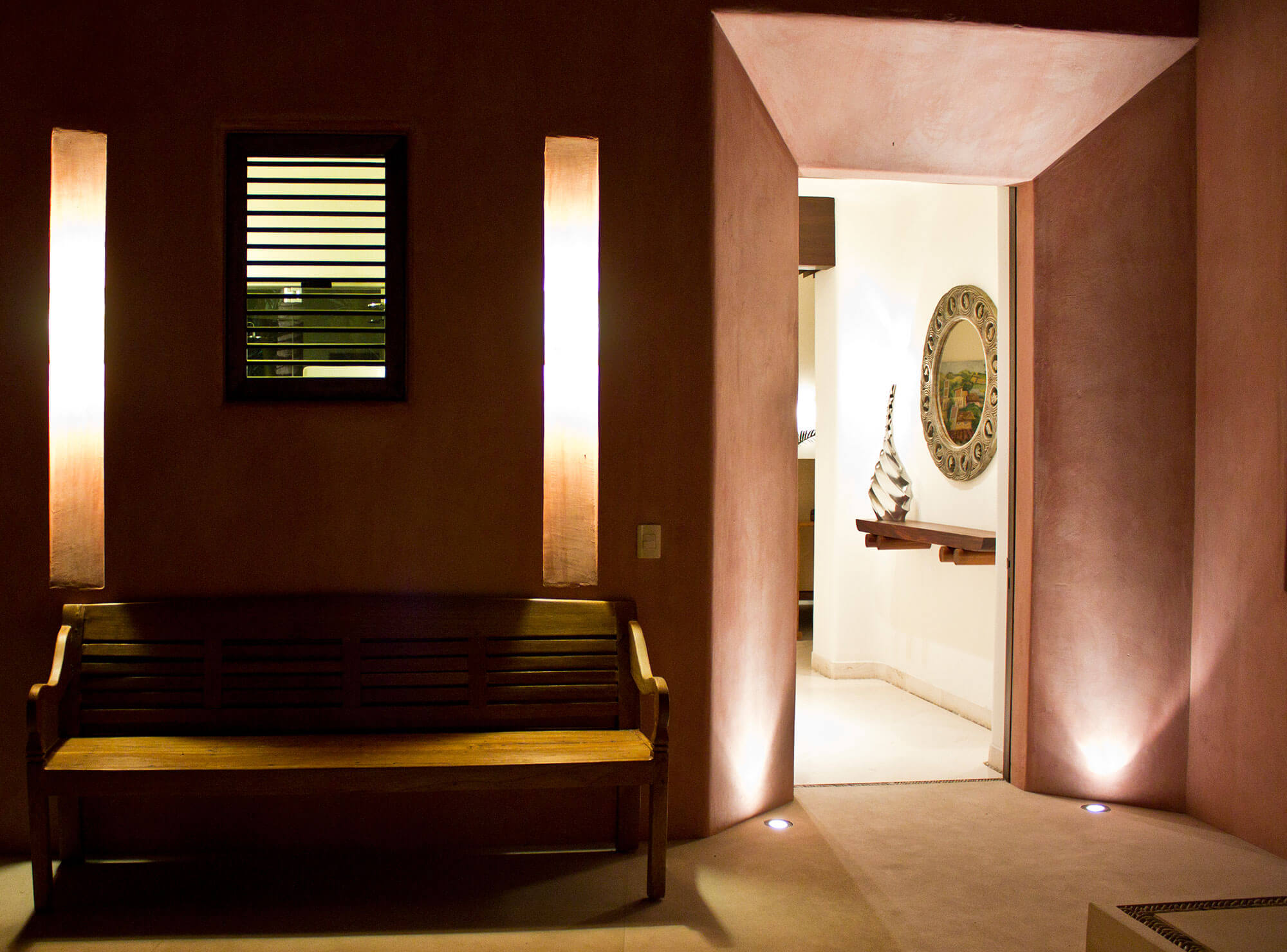 Arrival vestibule at night — terracotta plaster walls with paired vertical alabaster sconces flanking a louvered window, heavy teak bench against the wall, floor uplights, and an open archway revealing the hallway with ornate mirror and sculptural vase, Estate Venado, Ranchos Estates, Punta Mita.
