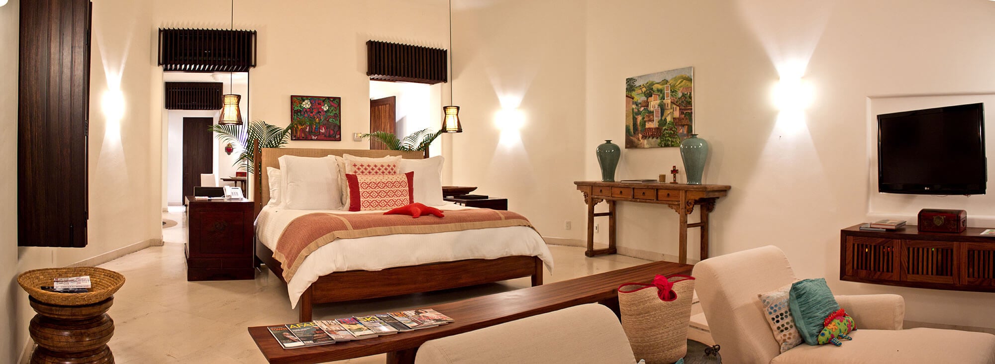 Wide bedroom interior with a king bed dressed in coral linen and red accent pillows, pendant lanterns, antique Chinese console with celadon vases, colorful oil painting, lounge seating with coffee table and travel magazines, and a flat-screen TV, Estate Venado, Ranchos Estates, Punta Mita.