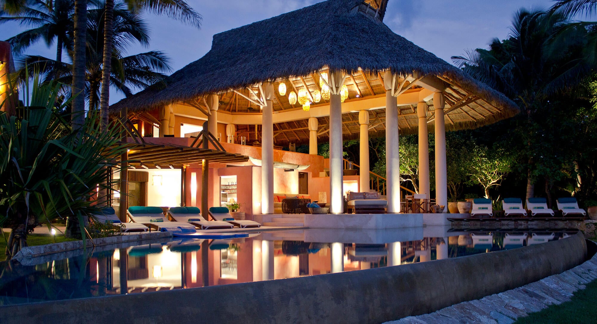 Full estate reflected in a still pool at dusk — the circular thatched palapa pavilion with pendant lanterns, white loungers with teal cushions, multi-level terracotta structure, and tall palms all mirrored in the water, Estate Venado, Ranchos Estates, Punta Mita.