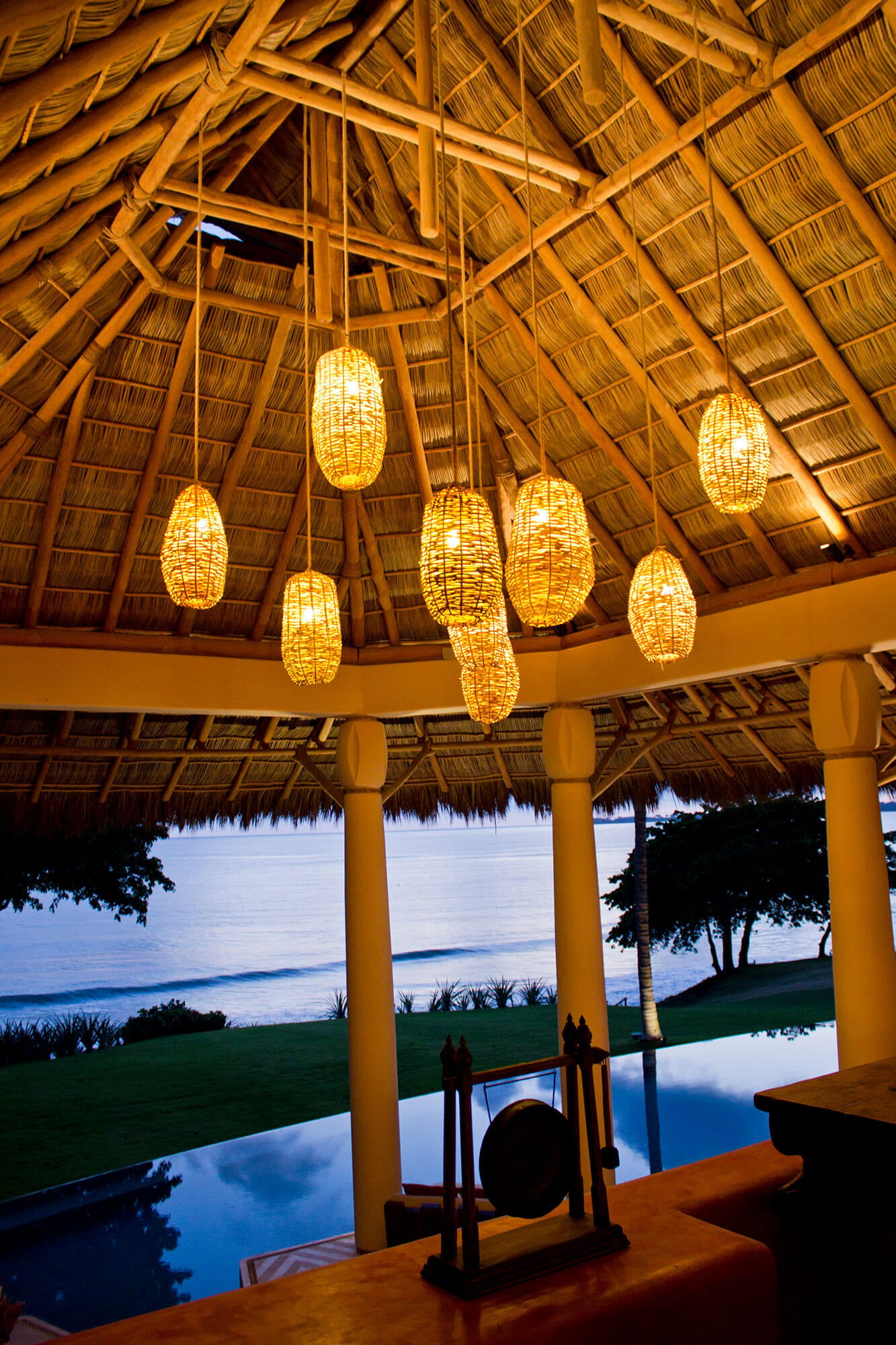 Looking up through the palapa ceiling at a cluster of woven rattan pendant lanterns glowing gold against thatched bamboo-and-palm-leaf rafters, with round stucco columns and a Pacific ocean view at dusk through the open sides, Estate Venado, Ranchos Estates, Punta Mita.