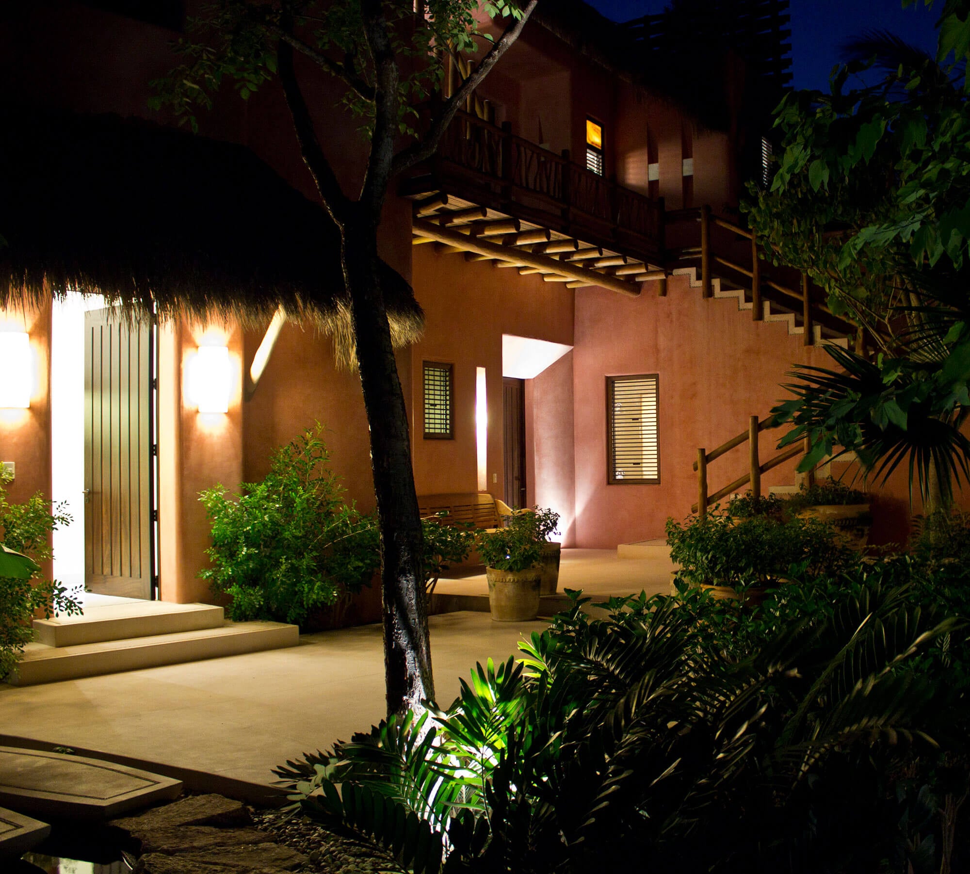 Night courtyard exterior — two-story terracotta estate facade with an external wooden staircase rising to an upper balcony, garden uplights illuminating a mature tree and lush tropical plantings, and a palapa-covered entry with wall sconces, Estate Venado, Ranchos Estates, Punta Mita.