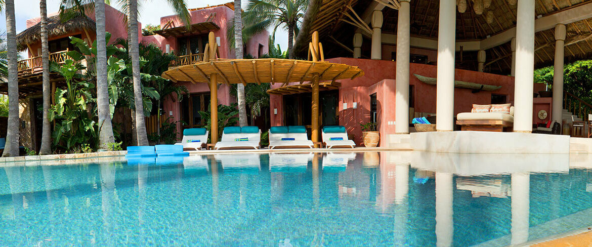 Daytime pool with clear turquoise water, white teak loungers fitted with teal cushions and blue foam floats, a rustic bamboo umbrella structure poolside, and the terracotta estate building with palapa pavilion and palms behind, Estate Venado, Ranchos Estates, Punta Mita.