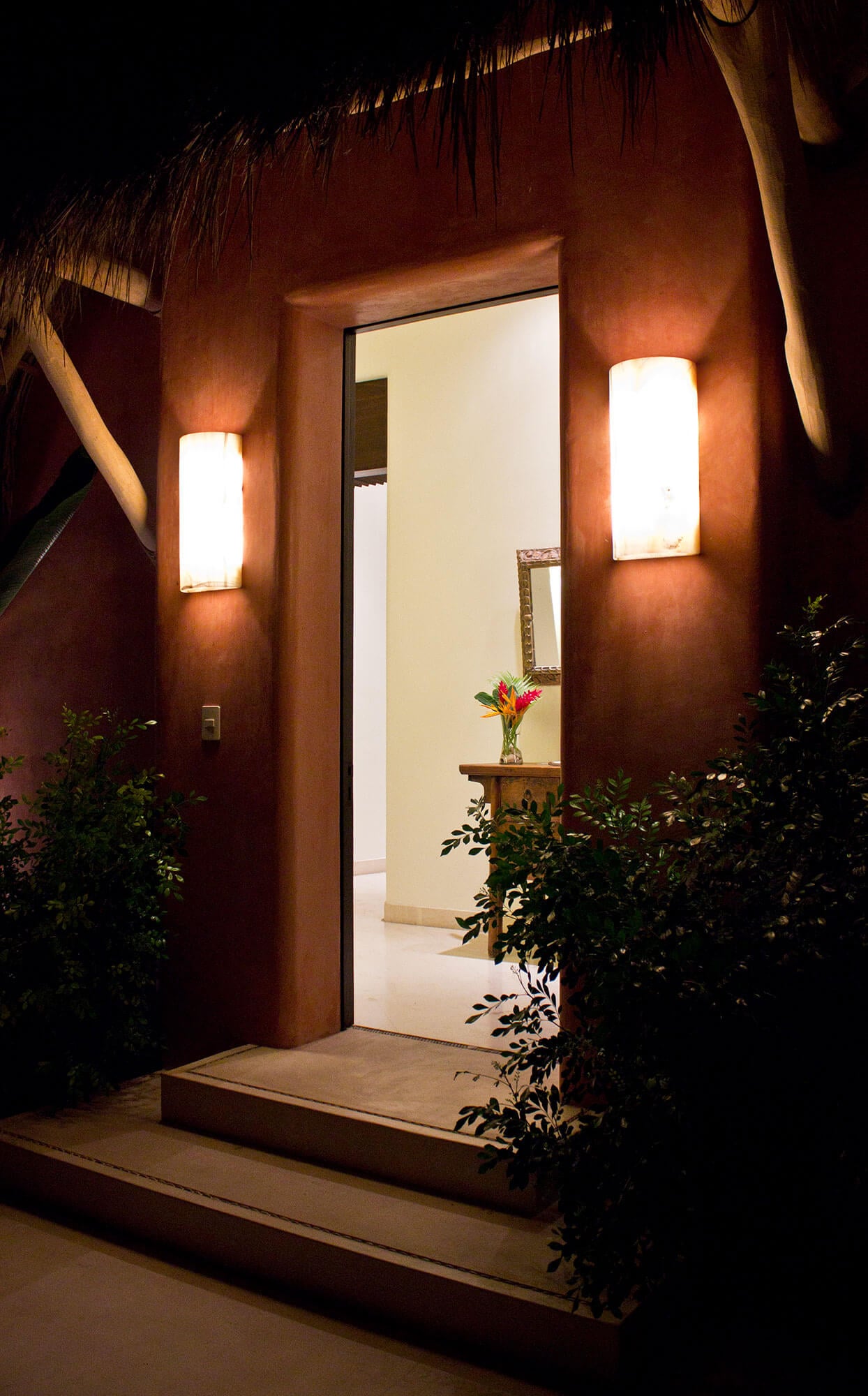 Night exterior entry door flanked by two alabaster cylinder sconces set into terracotta stucco, open door revealing a lit foyer with stone console table, tropical flower arrangement, and ornate mirror, framed by tropical shrubs and thatch above, Estate Venado, Ranchos Estates, Punta Mita.