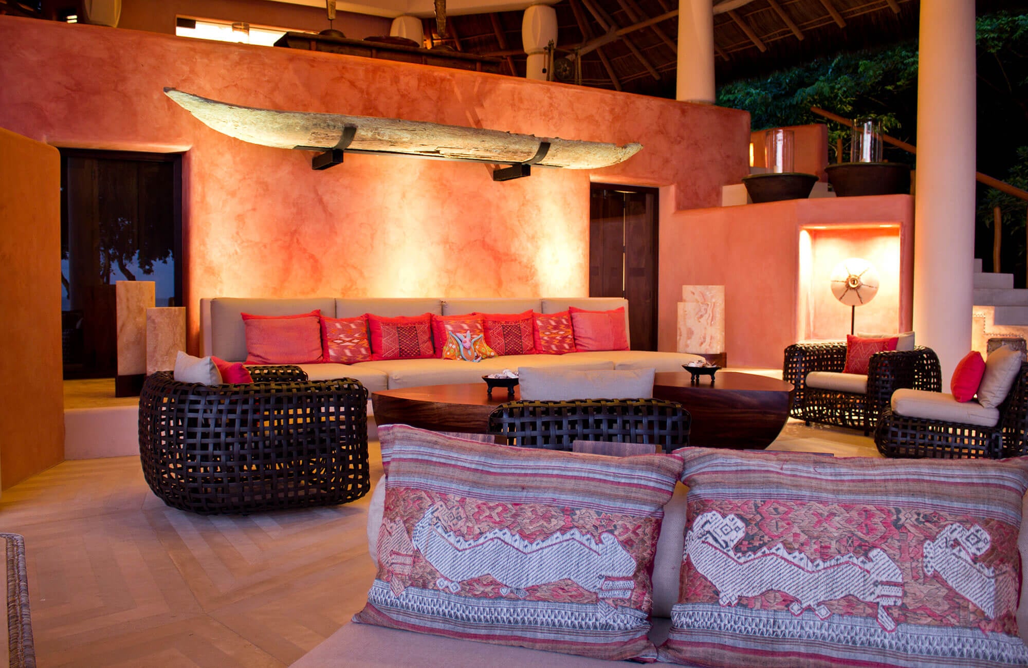 Living pavilion interior with a terracotta accent wall mounting an antique dugout canoe as sculpture, built-in curved sofa stacked with red and coral pillows, woven dark rattan armchairs, tribal textile cushions in the foreground, illuminated stone niches, and stucco columns, Estate Venado, Ranchos Estates, Punta Mita.
