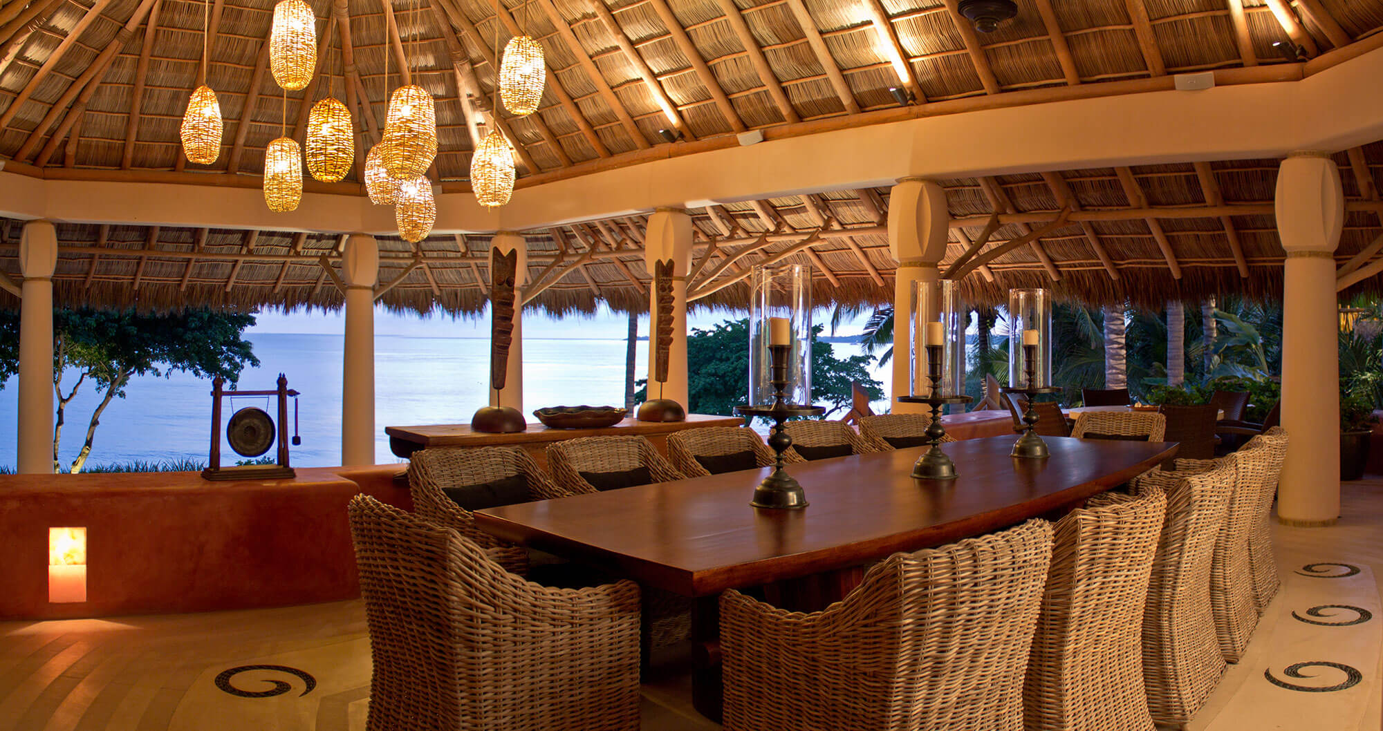 Open-air dining under the palapa with a long reclaimed-wood table seating ten in natural wicker chairs, cluster of woven rattan pendant lanterns above, brass candlestick centerpiece, and a framed Pacific ocean view through round stucco columns at dusk, Estate Venado, Ranchos Estates, Punta Mita.