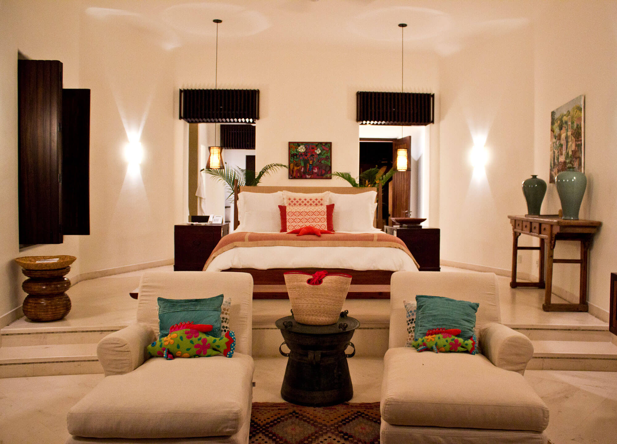 Suite bedroom with king bed on a raised marble platform, dark wood headboard, coral linen, pendant lanterns, colorful floral painting above, cream chaise longues with teal and embroidered pillows flanking a bronze drum table, and a celadon-vase console against the wall, Estate Venado, Ranchos Estates, Punta Mita.