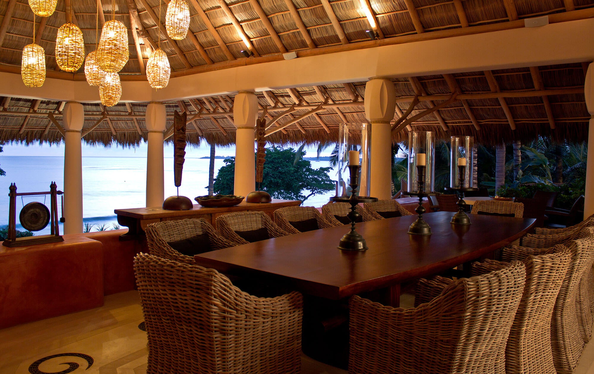 Palapa dining from the gong side — long wood dining table with wicker chairs, woven pendant lanterns overhead, a bronze gong on a stand at far left, brass candlestick centerpiece, and the ocean and palms visible through open round columns, Estate Venado, Ranchos Estates, Punta Mita.