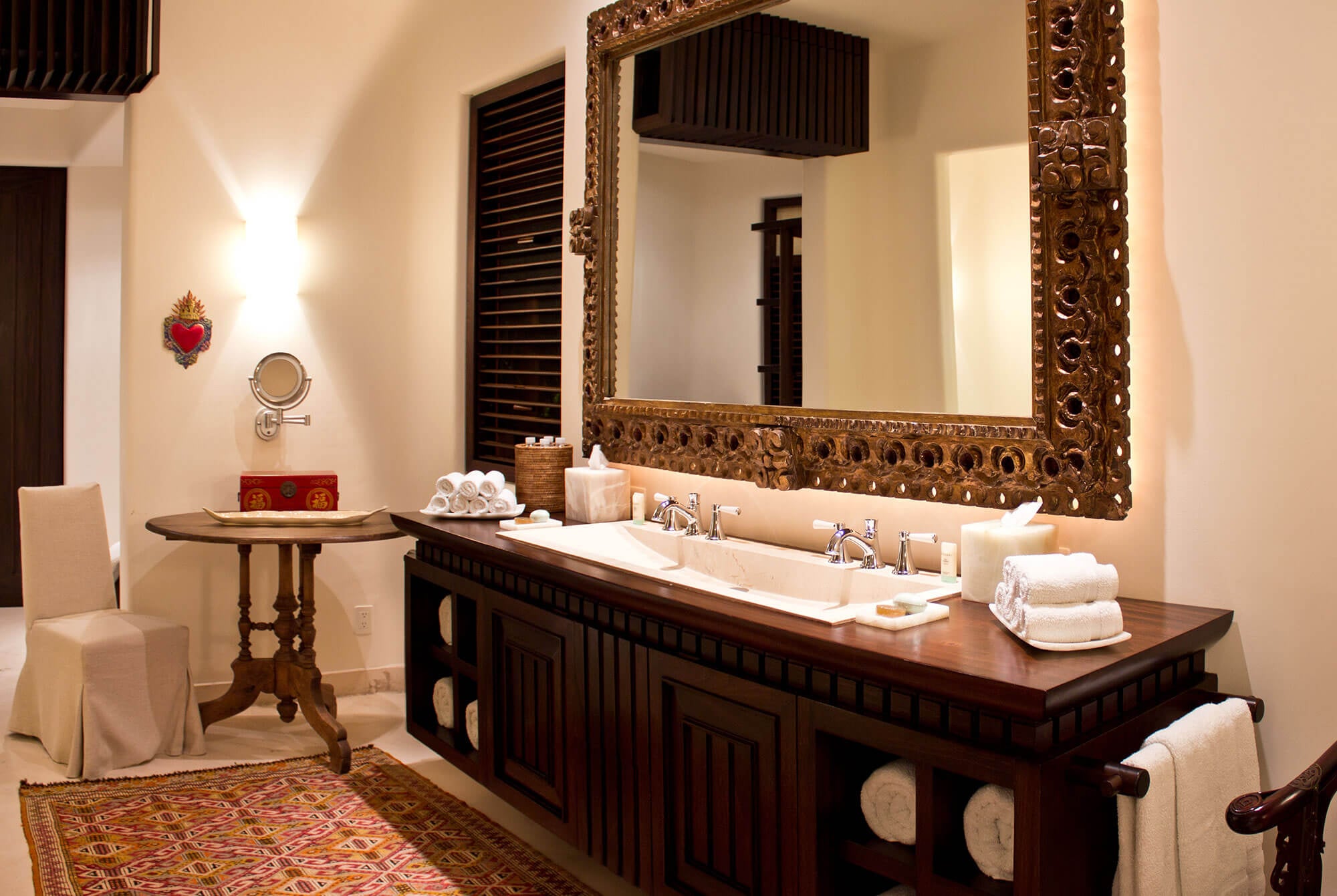 Ensuite bathroom with a double-sink dark wood vanity, carved gilt baroque mirror, rolled white towels, chrome fixtures, Oaxacan textile rug on marble floor, Mexican sacred-heart folk-art piece on the wall, and a round pedestal side table with lacquer box, Estate Venado, Ranchos Estates, Punta Mita.