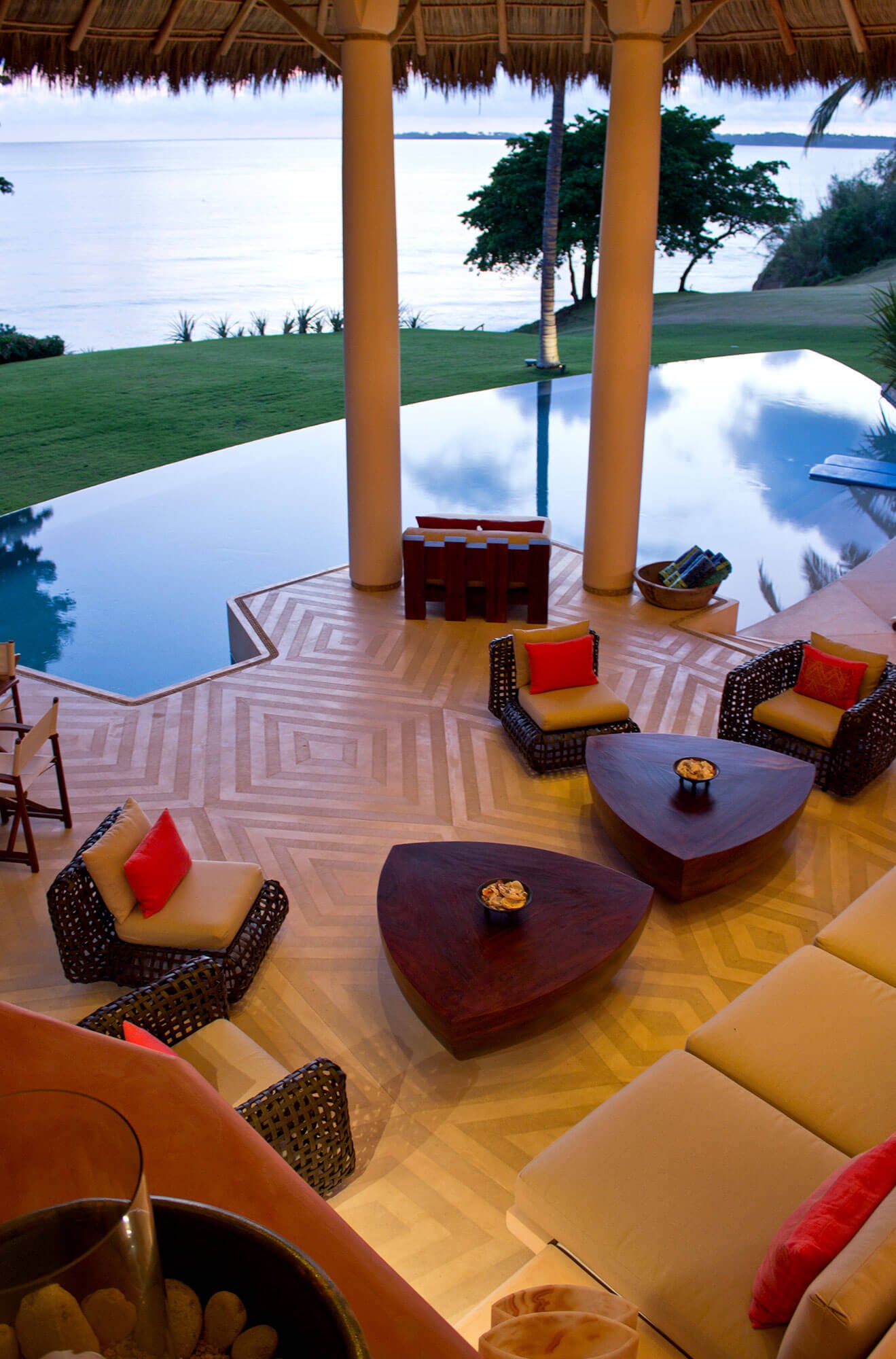 Overhead view of the open-air palapa living pavilion at dusk — wicker armchairs with orange cushions and triangular wood coffee tables arranged on an inlaid herringbone-stone floor, the still pool reflecting clouds beyond, Pacific ocean and lawn visible through round stucco columns, Estate Venado, Ranchos Estates, Punta Mita.