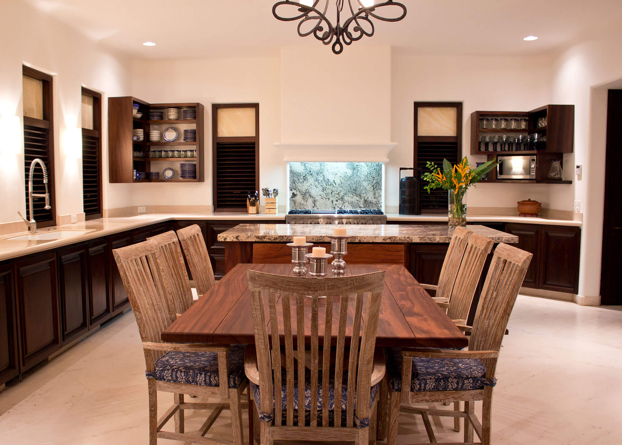 Spacious kitchen with dark mahogany cabinetry, a granite island backlit by a translucent stone panel, a wrought-iron chandelier overhead, and a rectangular wood dining table with six slatted chairs and indigo textile cushions on marble-tile floors, Estate Venado, Ranchos Estates, Punta Mita.