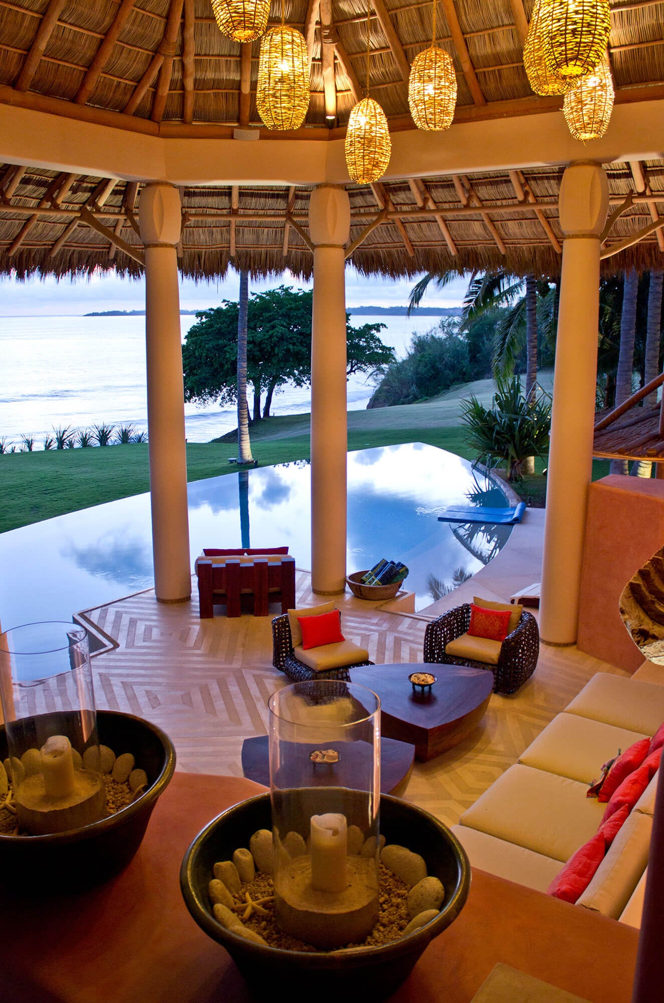 Inside the circular palapa at dusk — woven rattan pendant lanterns glow gold under the thatched canopy, wicker armchairs with orange cushions face triangular wood coffee tables on the inlaid floor, the still pool and Pacific beyond, Estate Venado, Ranchos Estates, Punta Mita.