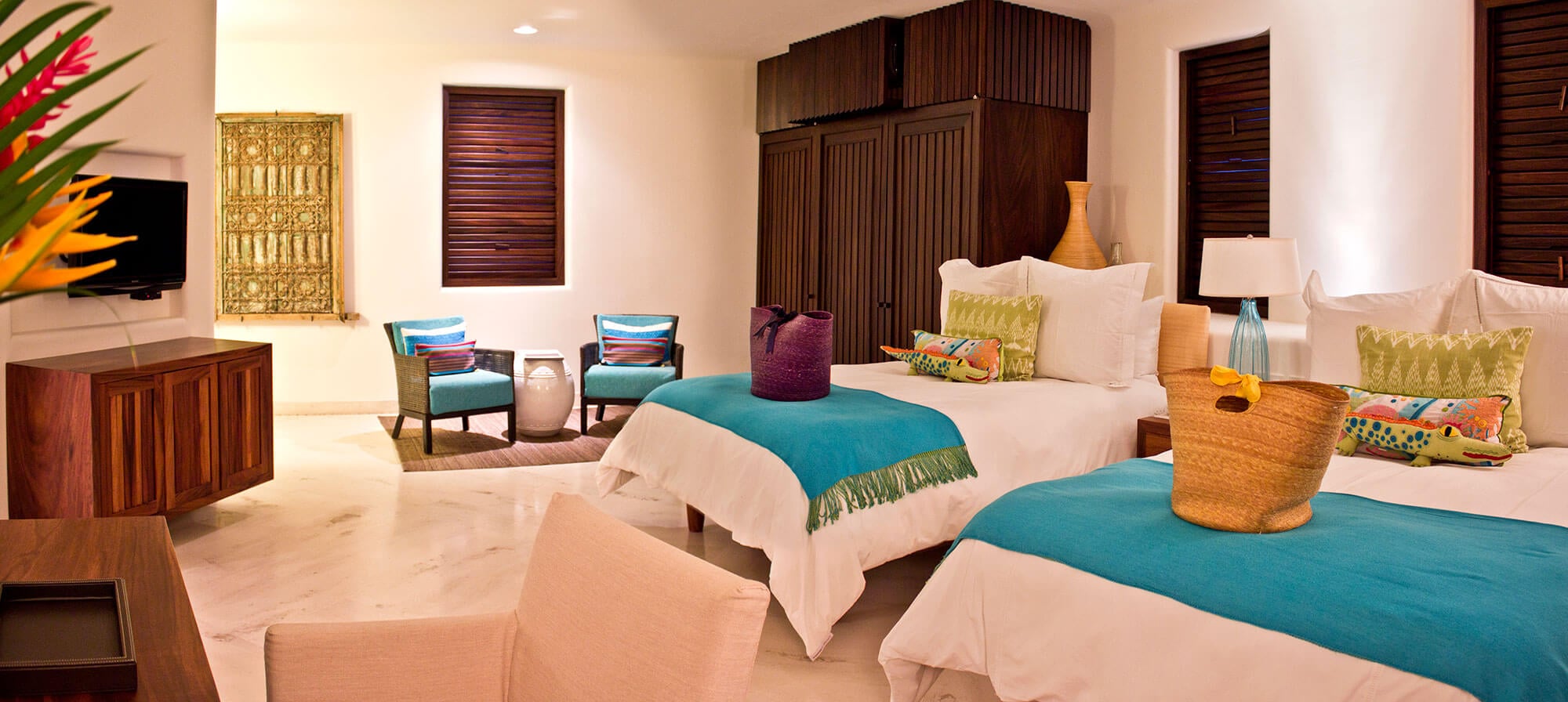 Twin-bed guest suite with two beds dressed in white linen and teal runners, a dark mahogany shuttered wardrobe, a gilded carved panel mounted on the wall beside the TV, two teal-upholstered side chairs with colorful embroidered pillows, and a straw market bag resting on one bed, Estate Venado, Ranchos Estates, Punta Mita.