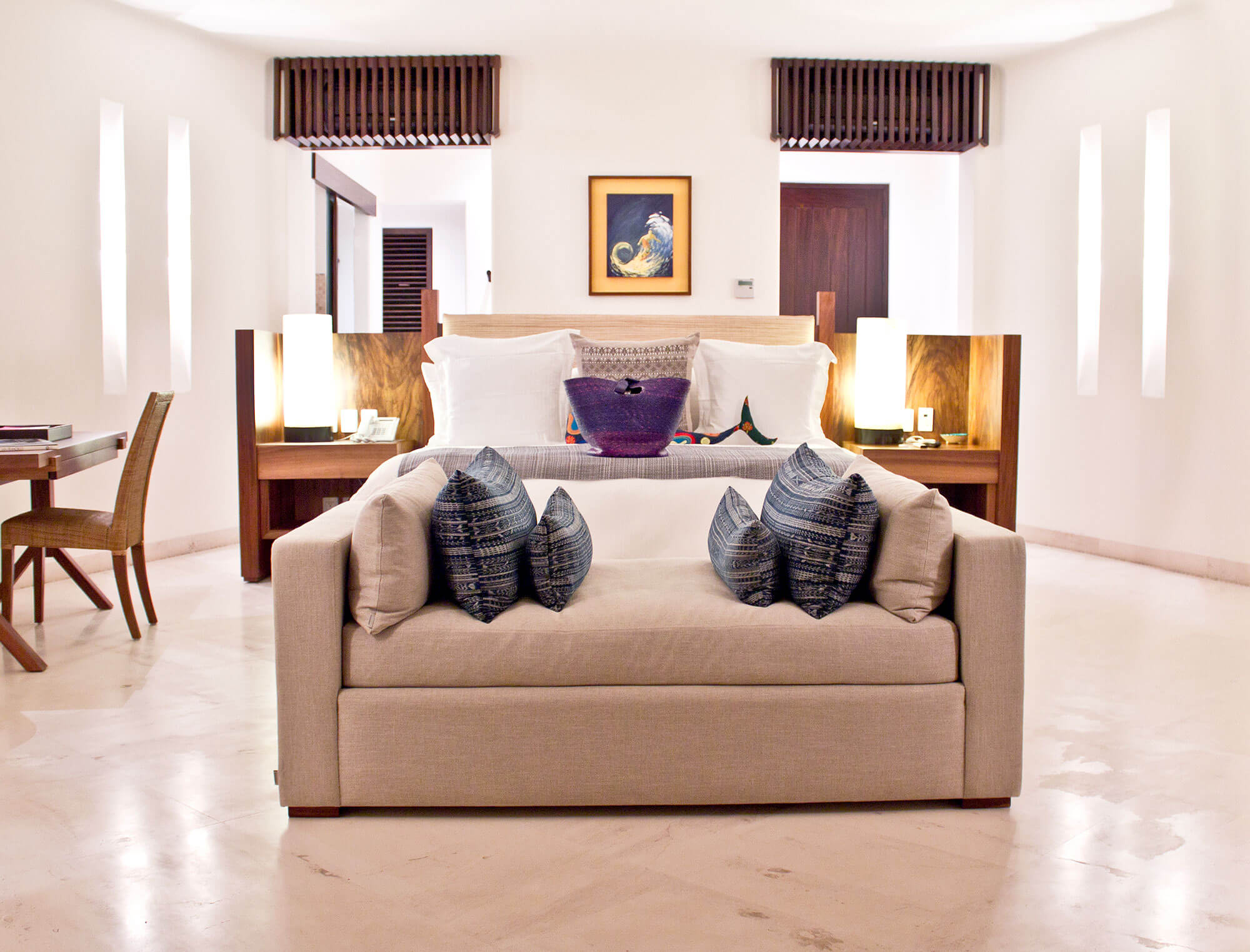 King bedroom with a wood-panel headboard integrating nightstand ledges and globe lamps, a framed wave painting above, a cream upholstered bench at the foot stacked with indigo ikat pillows, a purple woven basket on the bed, dark slatted valances over windows, and polished marble floors, Estate Venado, Ranchos Estates, Punta Mita.