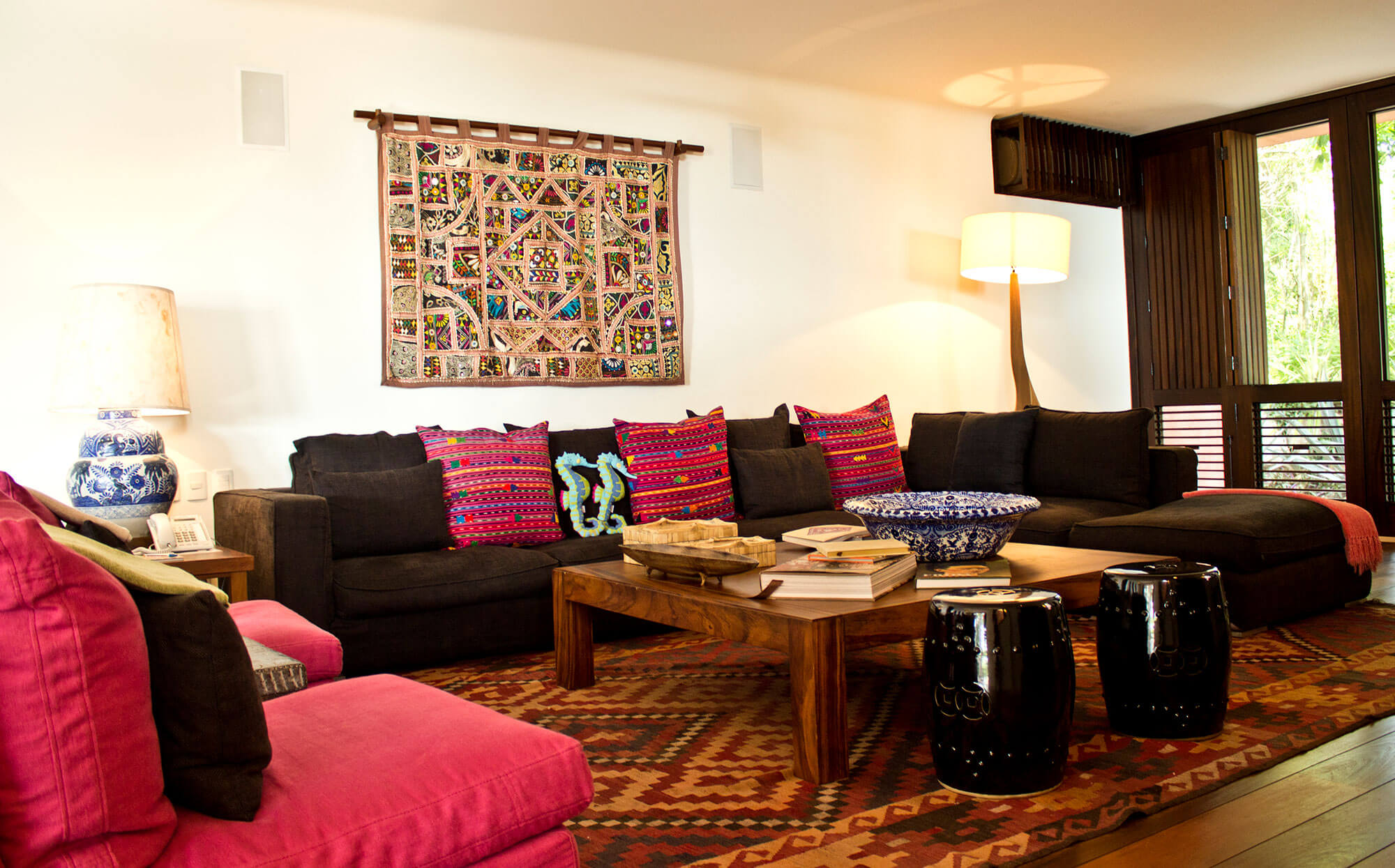 Interior sitting room with charcoal sofas piled with embroidered Mexican folk-art pillows, a kilim rug, a wood coffee table holding books and a blue Talavera bowl, an embroidered textile wall hanging on a bamboo rod, black Chinese garden stools, and dark louvered shutters at the window, Estate Venado, Ranchos Estates, Punta Mita.