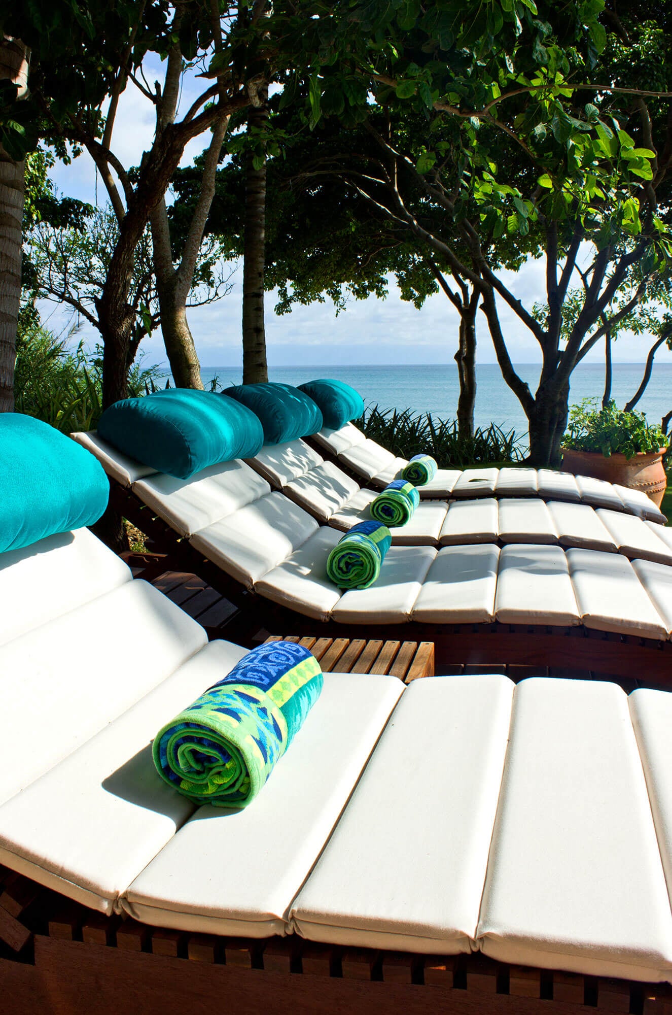 A row of five teak sun loungers with white cushions, teal bolster pillows, and a rolled green towel on each, set in dappled shade under mature trees with the Pacific visible through the foliage, Estate Venado, Ranchos Estates, Punta Mita.