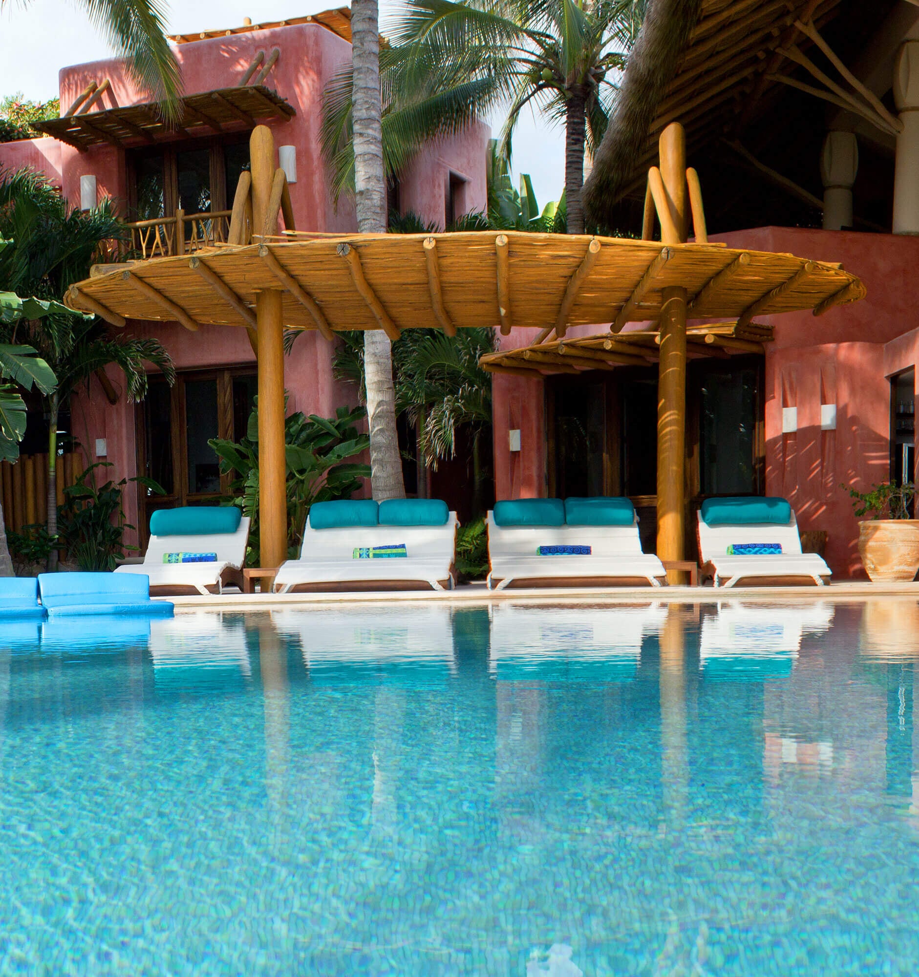 Pool viewed from the water's edge — white teak loungers with teal cushions line up under a large round log-and-bamboo shade structure, the terracotta estate building rising two storeys behind with a balcony, tall palms framing the scene on a clear afternoon, Estate Venado, Ranchos Estates, Punta Mita.