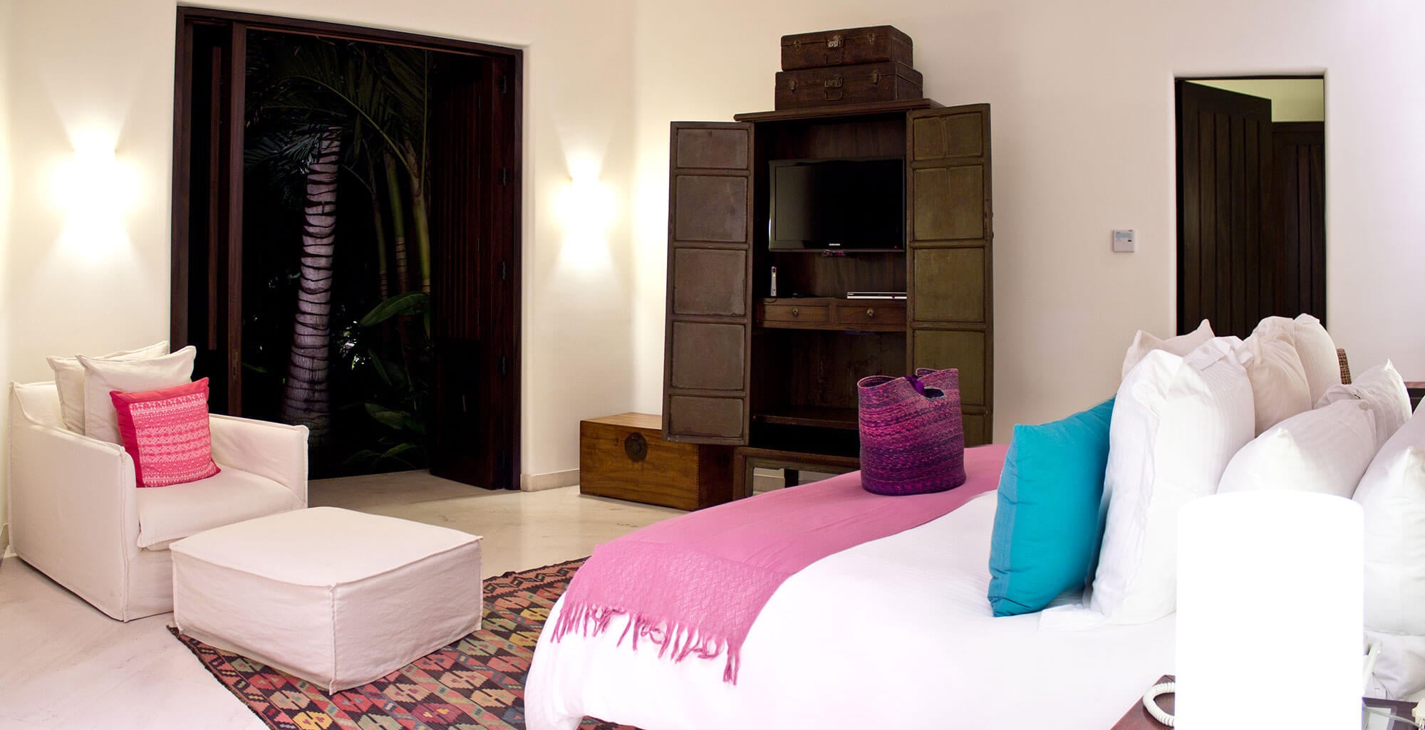 Guest bedroom with a king bed in white linen and a pink woven throw, a large distressed dark-wood armoire housing a flat-screen TV with vintage trunks stacked on top, a white upholstered armchair with a pink embroidered pillow and matching ottoman, a kilim rug underfoot, and open folding wood doors to a night garden, Estate Venado, Ranchos Estates, Punta Mita.