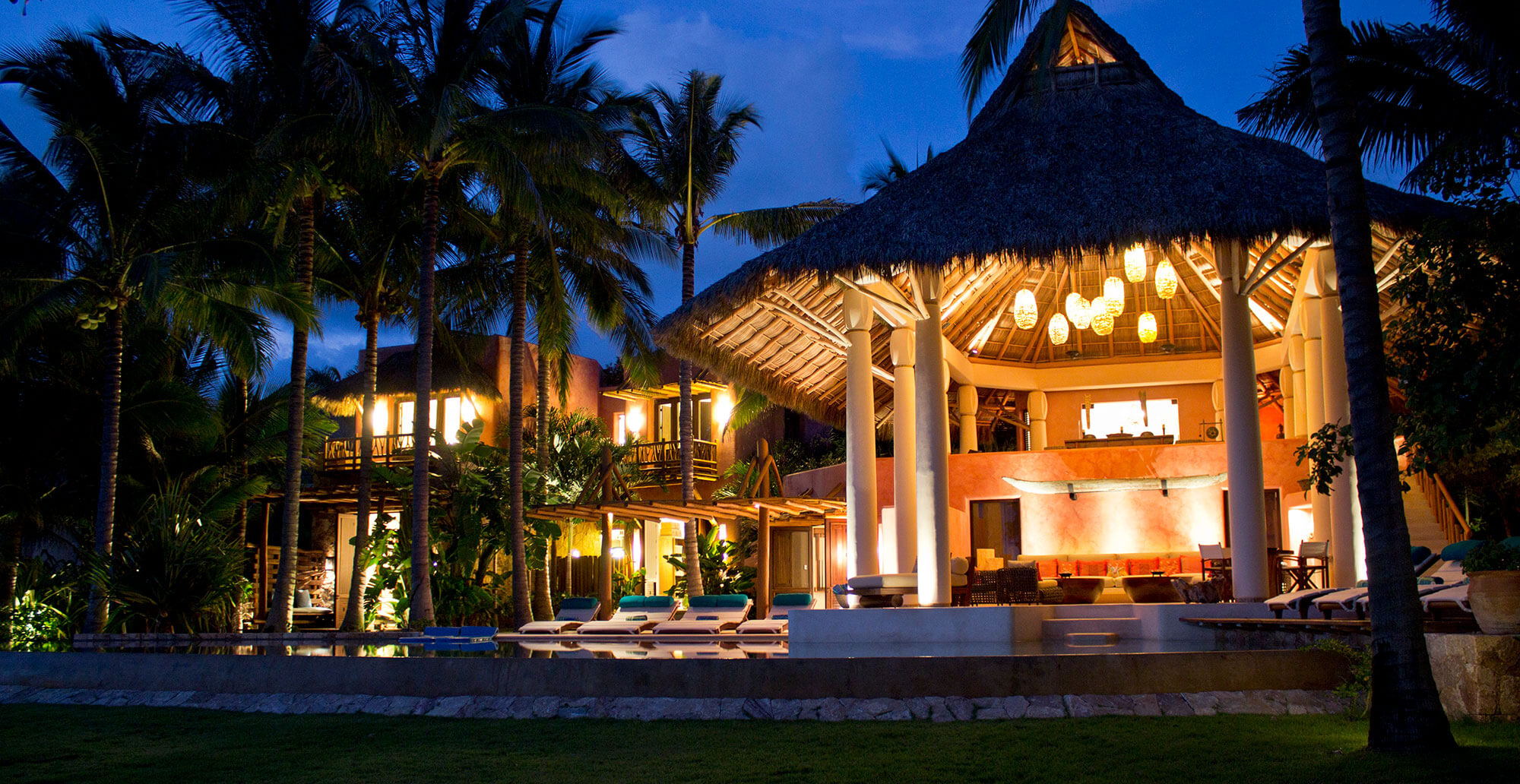 Night exterior of the full estate seen from the lawn — the circular thatched palapa pavilion lit with woven pendant lanterns, the multi-level terracotta building rising behind with lit balconies, pool and loungers in the foreground grass, tall palms silhouetted against the dark sky, Estate Venado, Ranchos Estates, Punta Mita.
