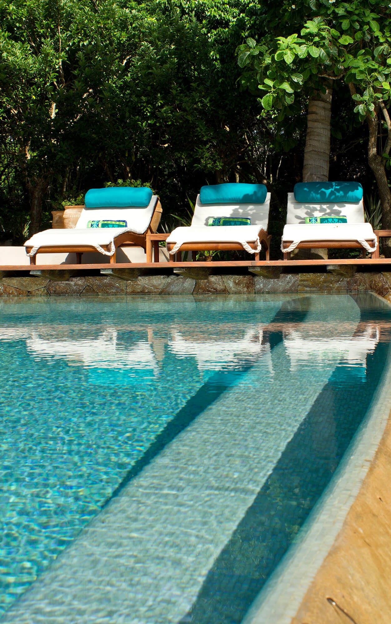 Three white teak loungers with teal head cushions and rolled green towels arranged beside the pool's stone coping, tropical garden trees reflected in the still water beyond, Estate Venado, Ranchos Estates, Punta Mita.