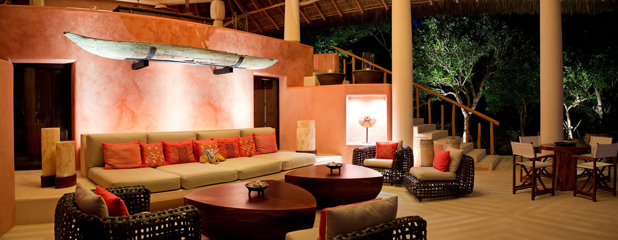 Palapa living pavilion at night from the side — a full-length antique dugout canoe mounted on the terracotta accent wall overhead, a wide built-in sofa with coral pillows at left, wicker armchairs flanking triangular wood coffee tables on the herringbone floor, a lit stone niche with folk-art figure, and an open staircase to the upper terrace at right, Estate Venado, Ranchos Estates, Punta Mita.