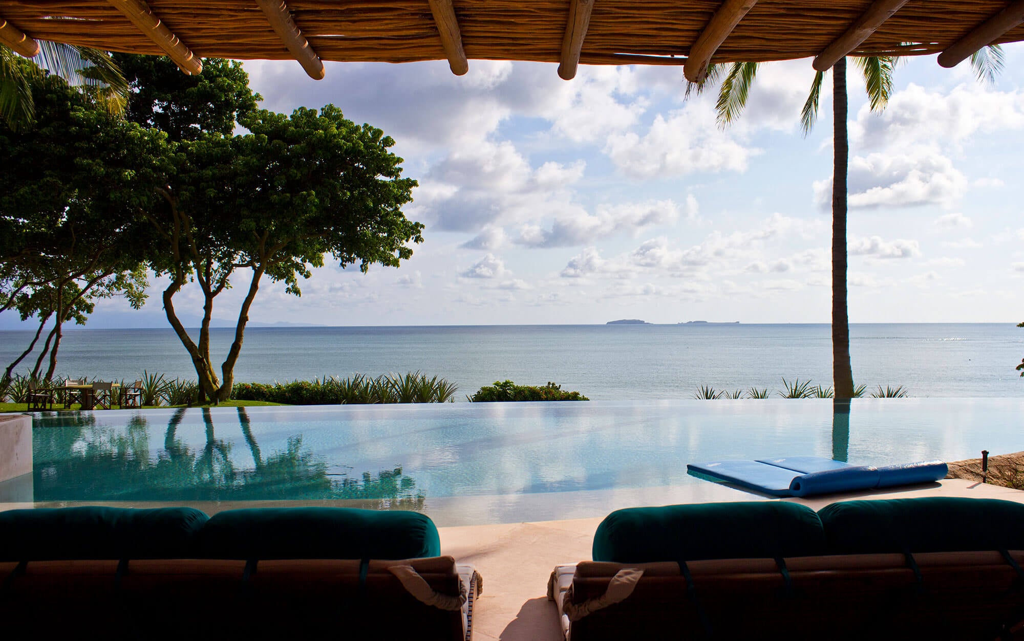 Looking out from under the palapa's log-and-bamboo ceiling across the infinity pool, where the water appears to dissolve into the Pacific horizon — Marietas Islands visible in the distance, a blue foam float on the pool ledge, teal-cushioned lounger headrests in the foreground, Estate Venado, Ranchos Estates, Punta Mita.