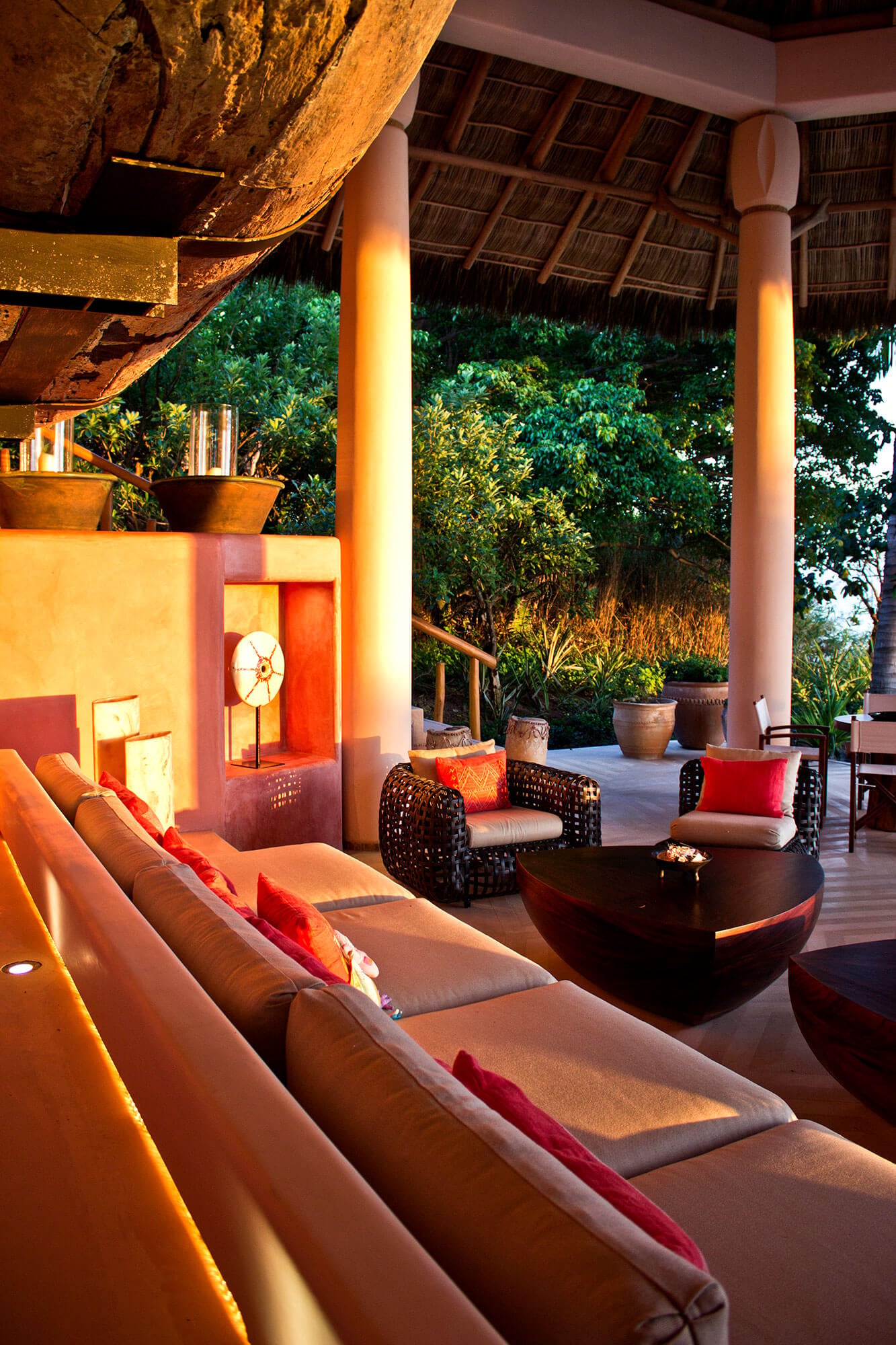 Evening view along the palapa sofa — the worn hull of the antique dugout canoe fills the top of frame above the curved terracotta wall, wicker armchairs and triangular coffee tables lead the eye toward the open columns framing the tropical garden in fading golden light, Estate Venado, Ranchos Estates, Punta Mita.