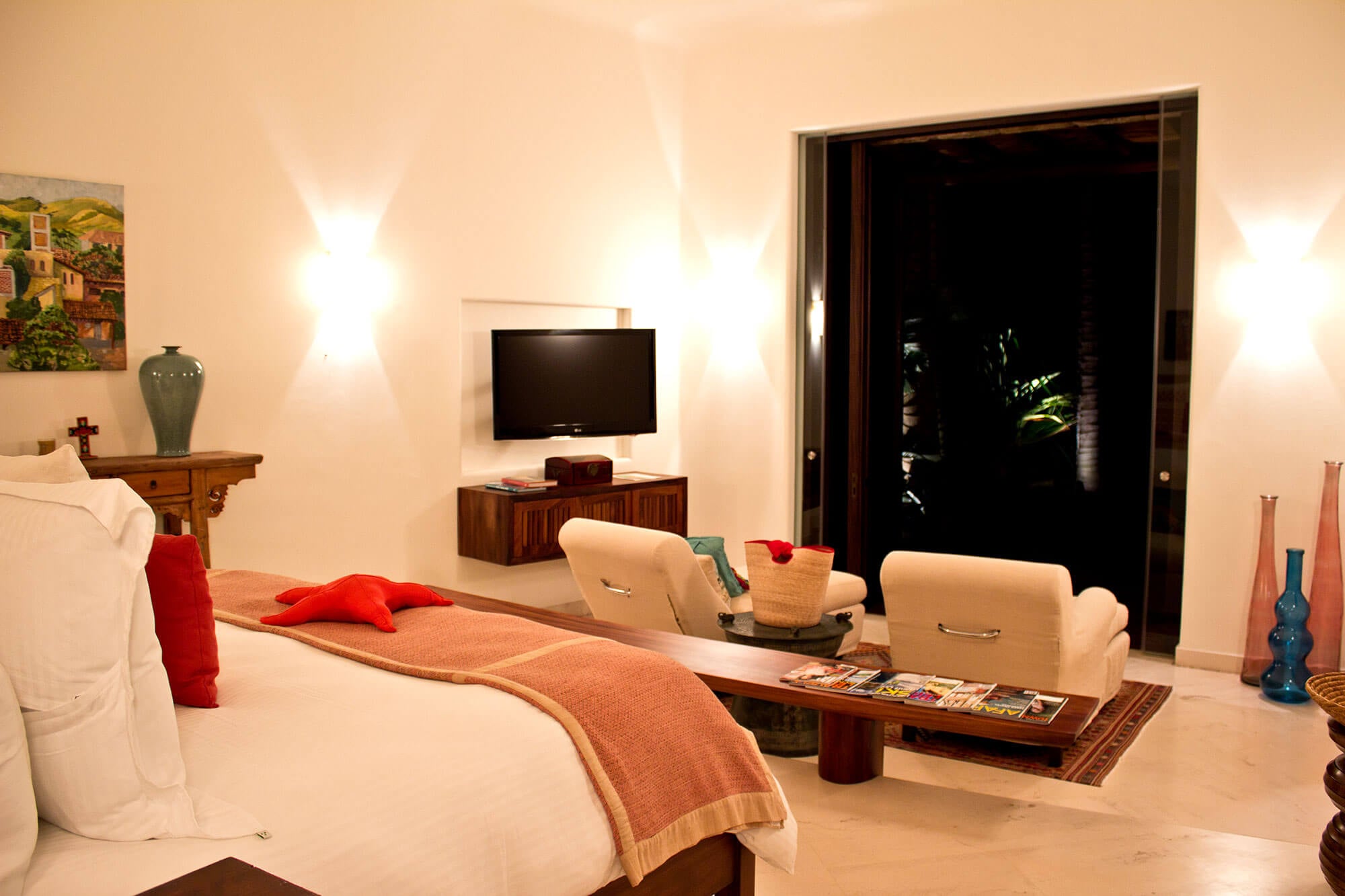 Guest bedroom at night — a king bed with white linen and a salmon woven runner, wall-mounted TV on a dark wood media console, two cream bucket chairs with travel magazines on a low coffee table, wall sconces, a celadon vase on an antique wood console, a colorful Mexican painting, glass floor vases, and a large sliding glass door to the dark garden, Estate Venado, Ranchos Estates, Punta Mita.
